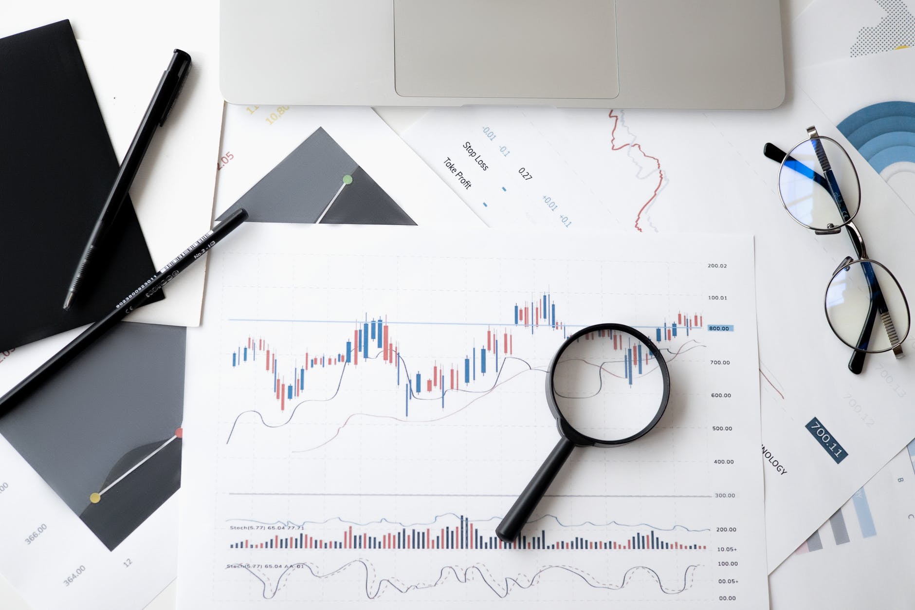 Desk with paper with graphs and magnifying glass on top of one part of line graph. A pair of pencils is on the left and glasses on the top right corner.
