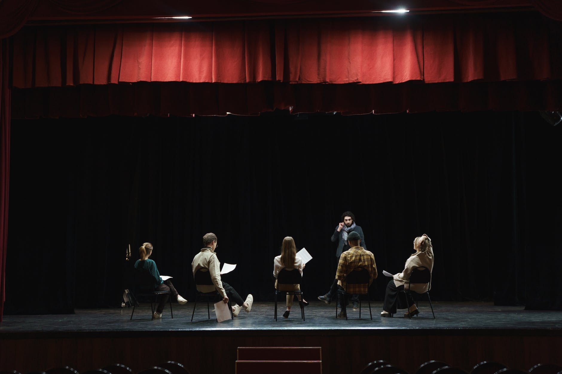 Five people seated on stage facing a man with black hair in the middle/right of the circle of chairs. Some of the people are holding paper scripts.