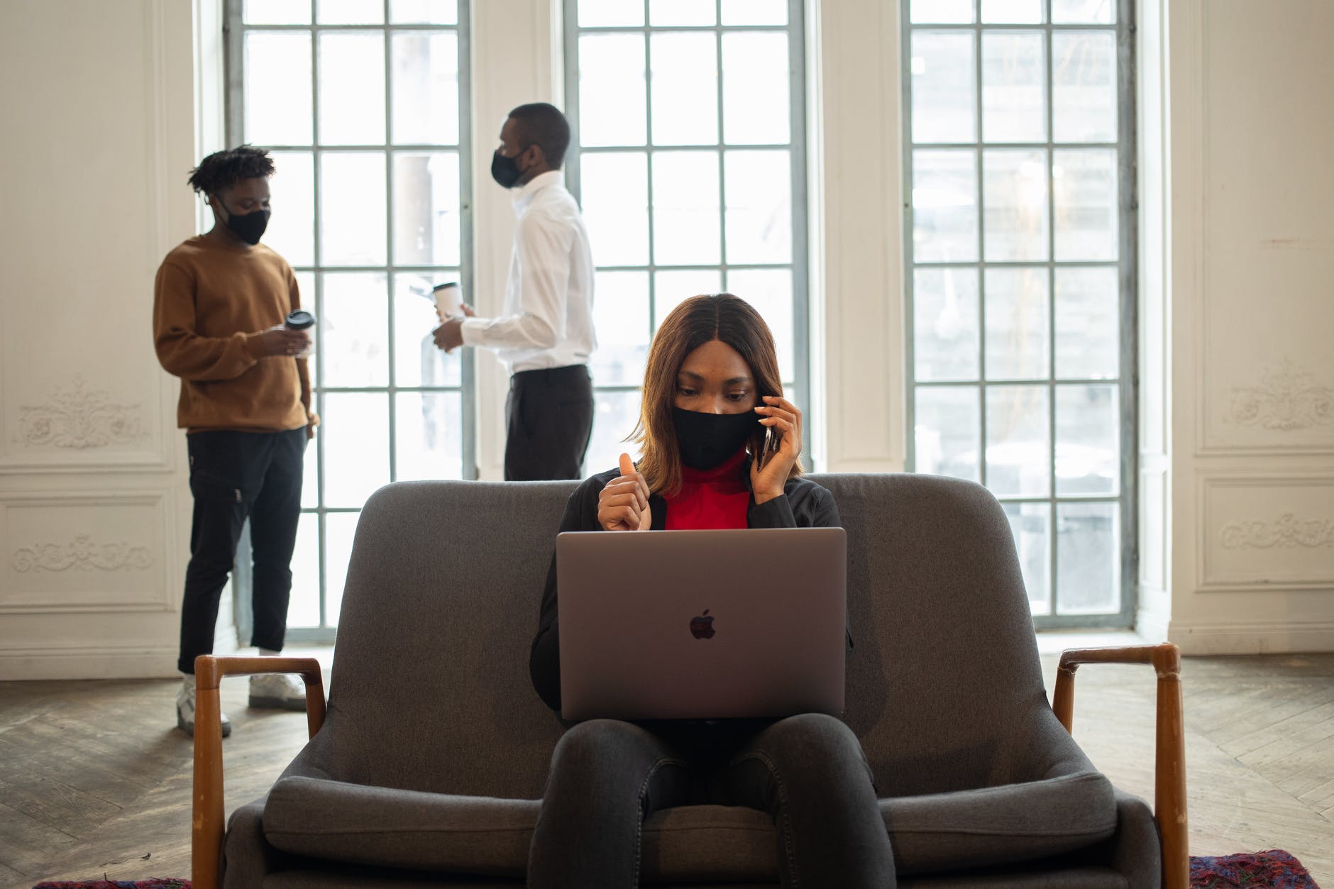 Black woman on phone with open laptop and wearing mask seated at couch. Behind two men holding coffee cups and wearing black masks are standing talking.
