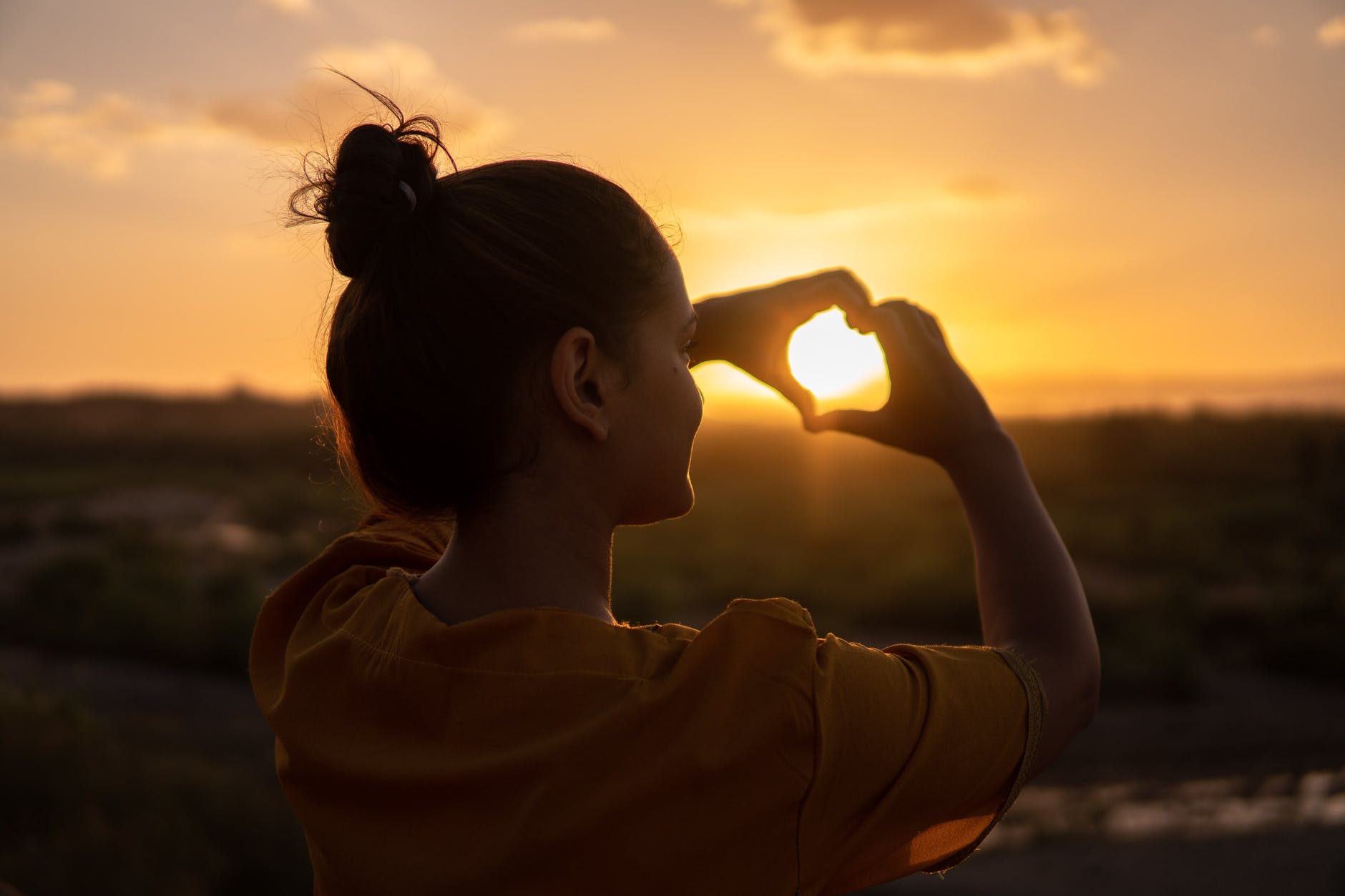 Woman facing sunset with hair in bun and making heart shape with hands and fingers.