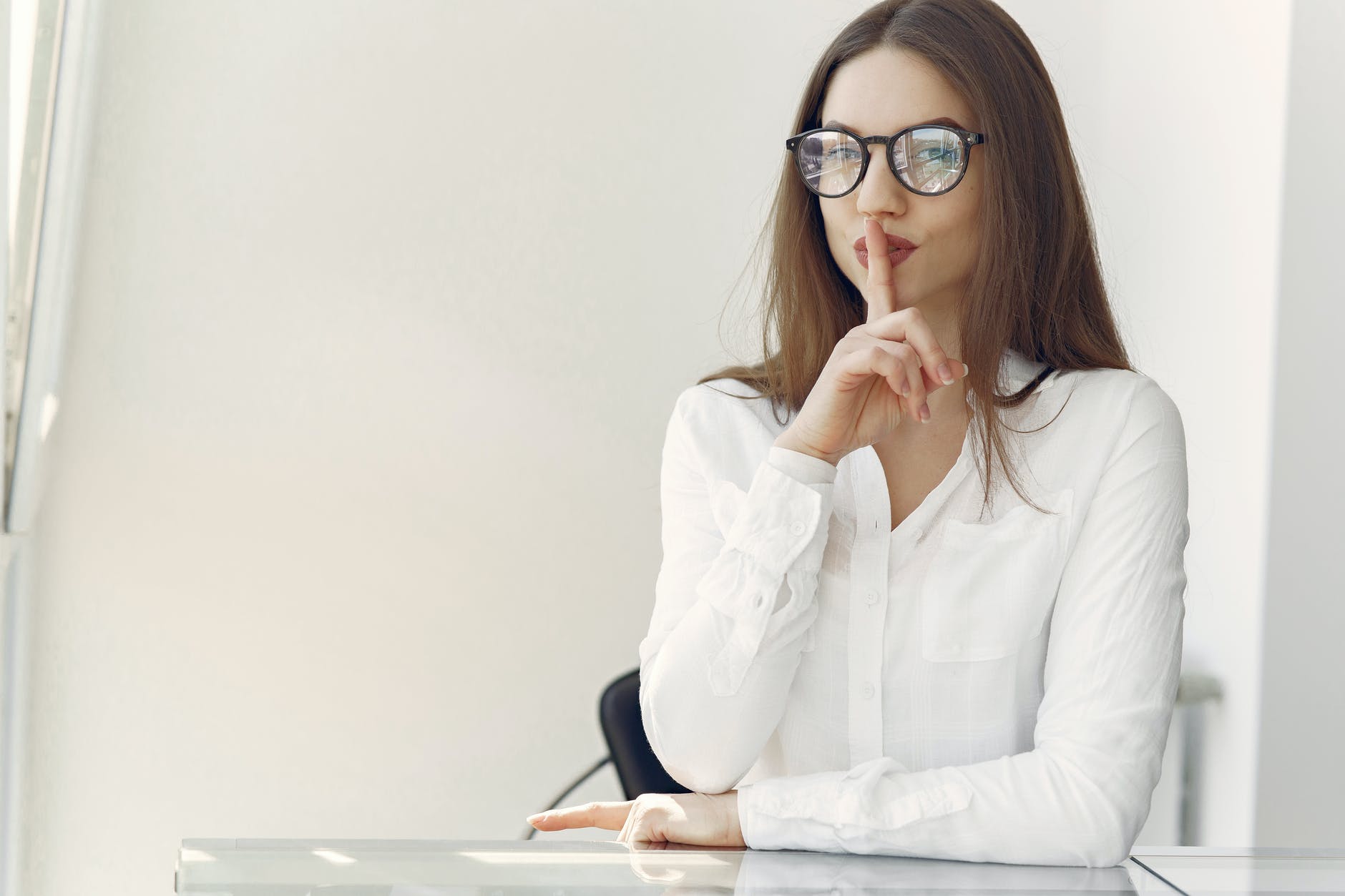 Woman with raised finger on lips and long brown hair and glasses.