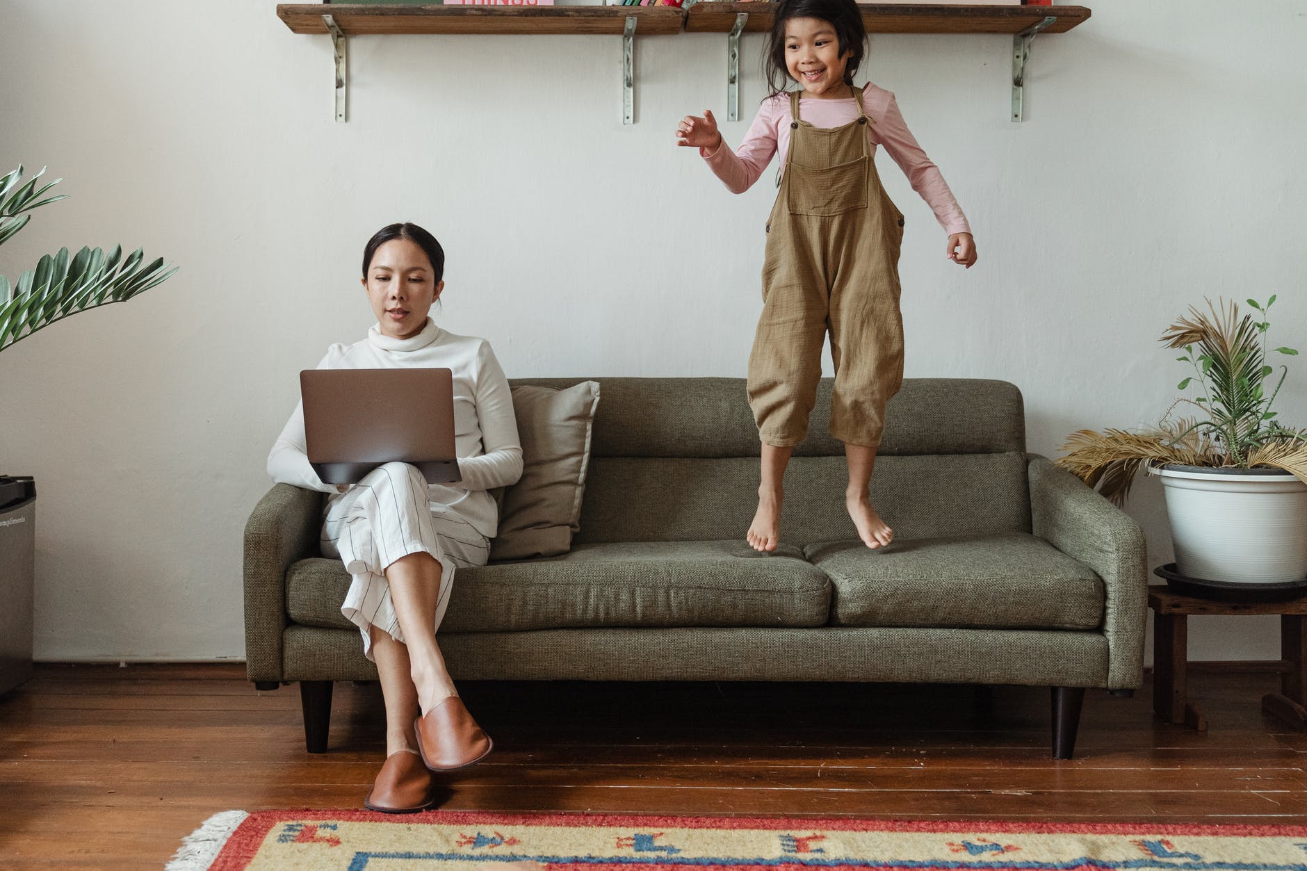 Woman working on laptop on one side of couch with child jumping on couch on the other side.