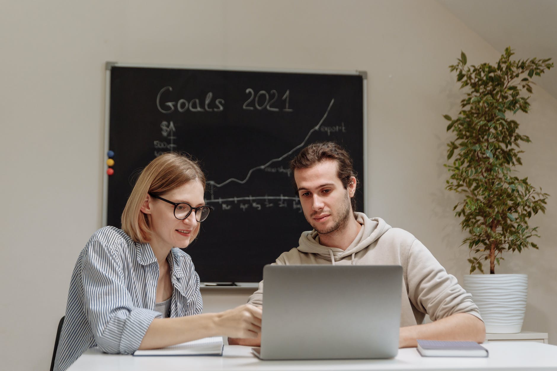Woman with glasses and shoulder-length hair pointing at laptop. Man is using laptop. Behind them, a black whiteboard has text "Goals 2021" and a graph with an upward line.