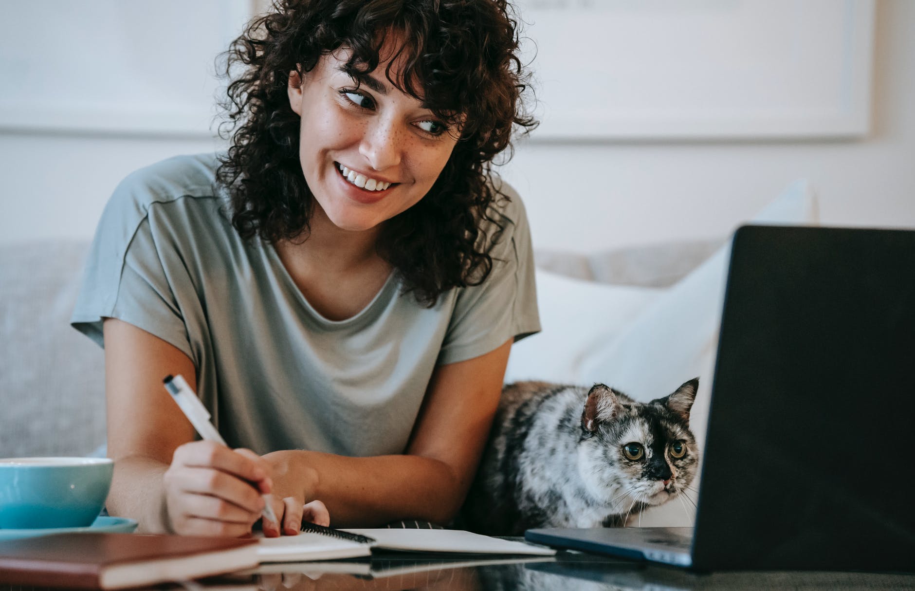 Woman smiling and writing next to cat and laptop. 