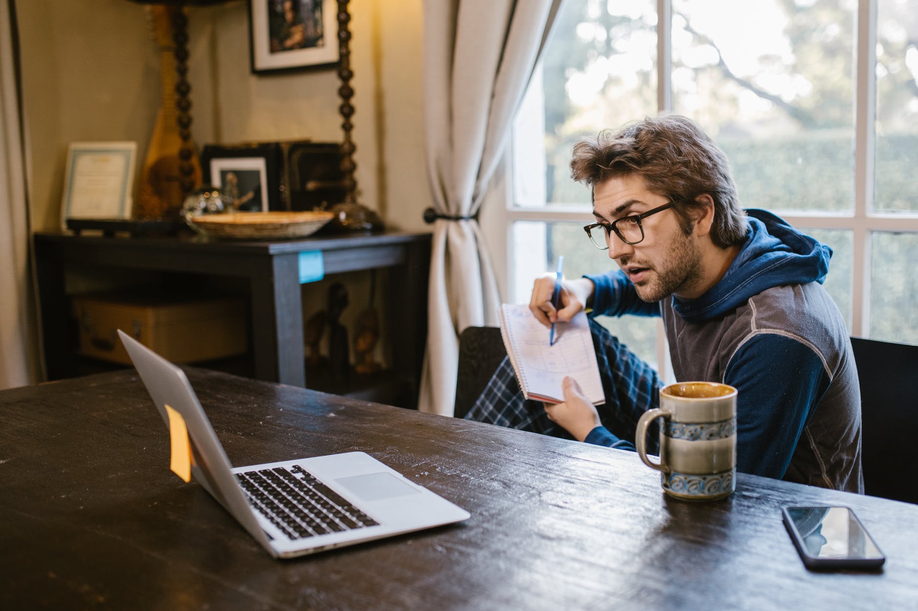 Person wearing hoodie and glasses taking notes with right hand and open laptop on desk.