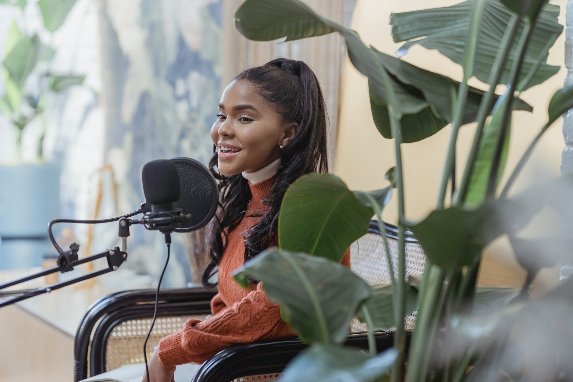 Black woman laughing/speaking into microphone with pop filter. Plants are out of focus.