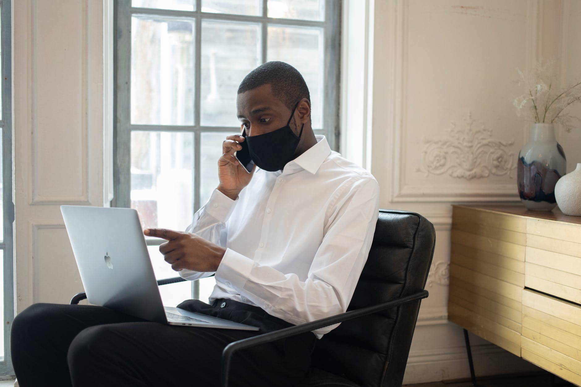 Black man wearing mask and white shirt. Laptop is on lab and man is talking to someone on the phone.