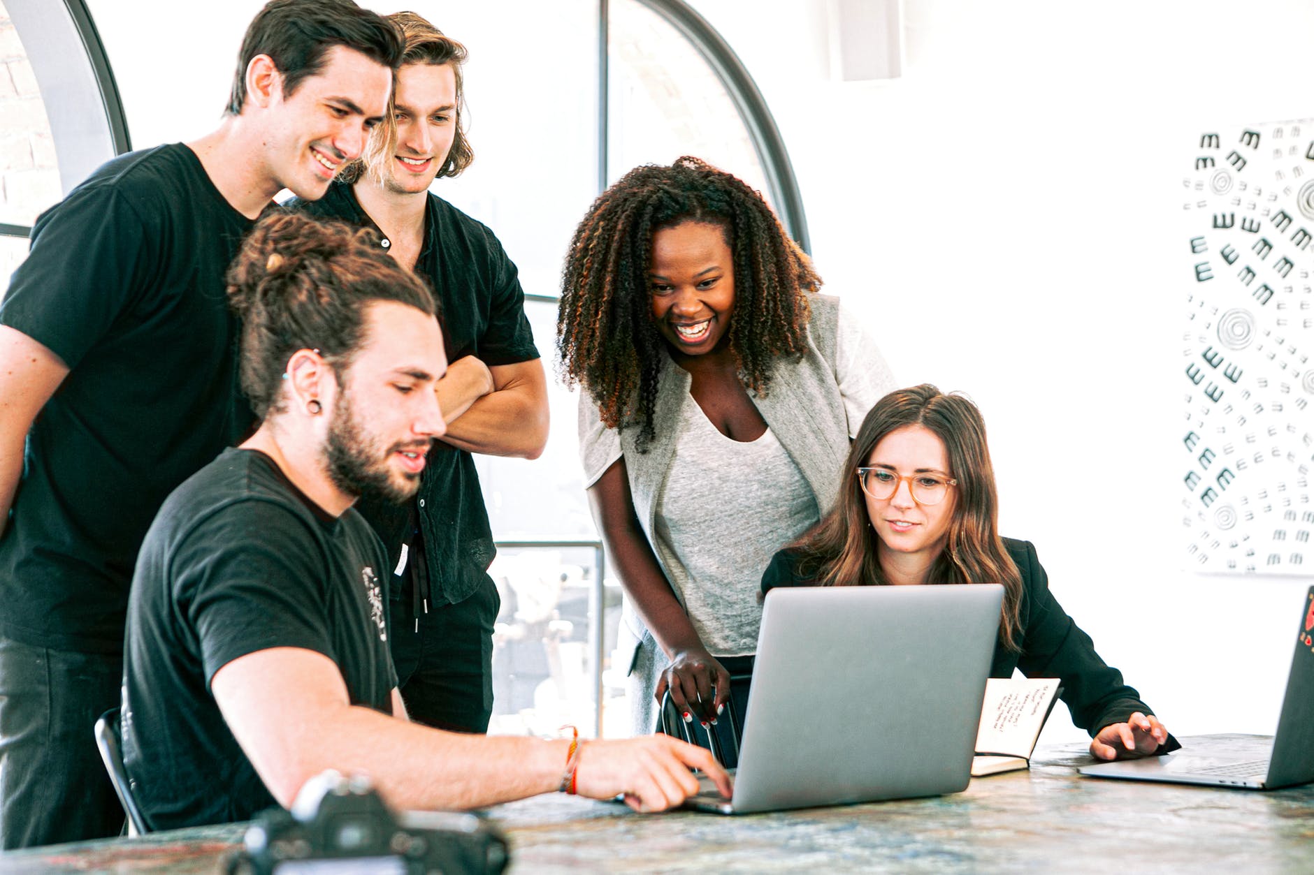 Five people around one laptop. Some are laughing and two are seated.