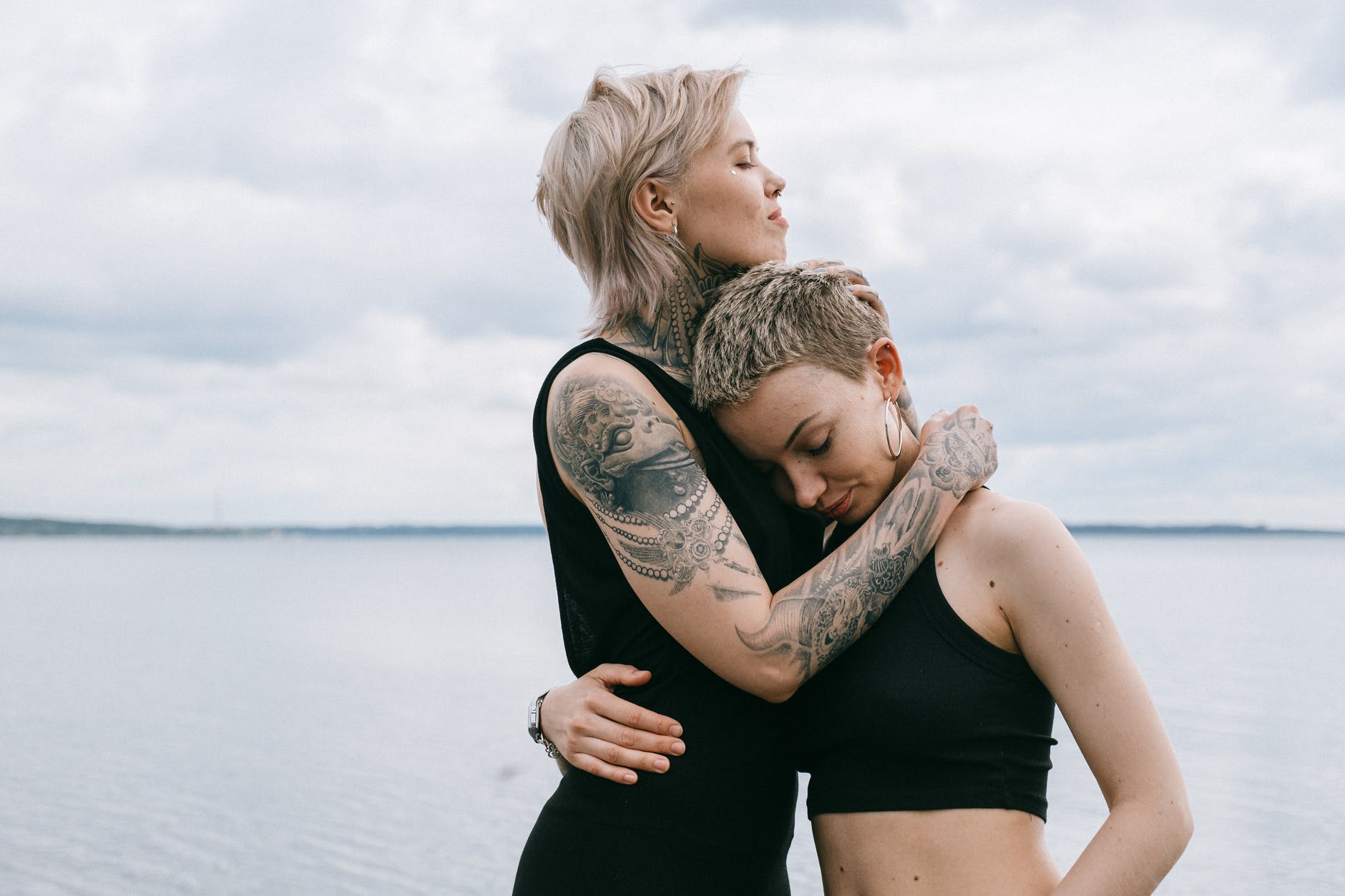 Two people embracing. Water is in the background. One person has tattoos on arm.