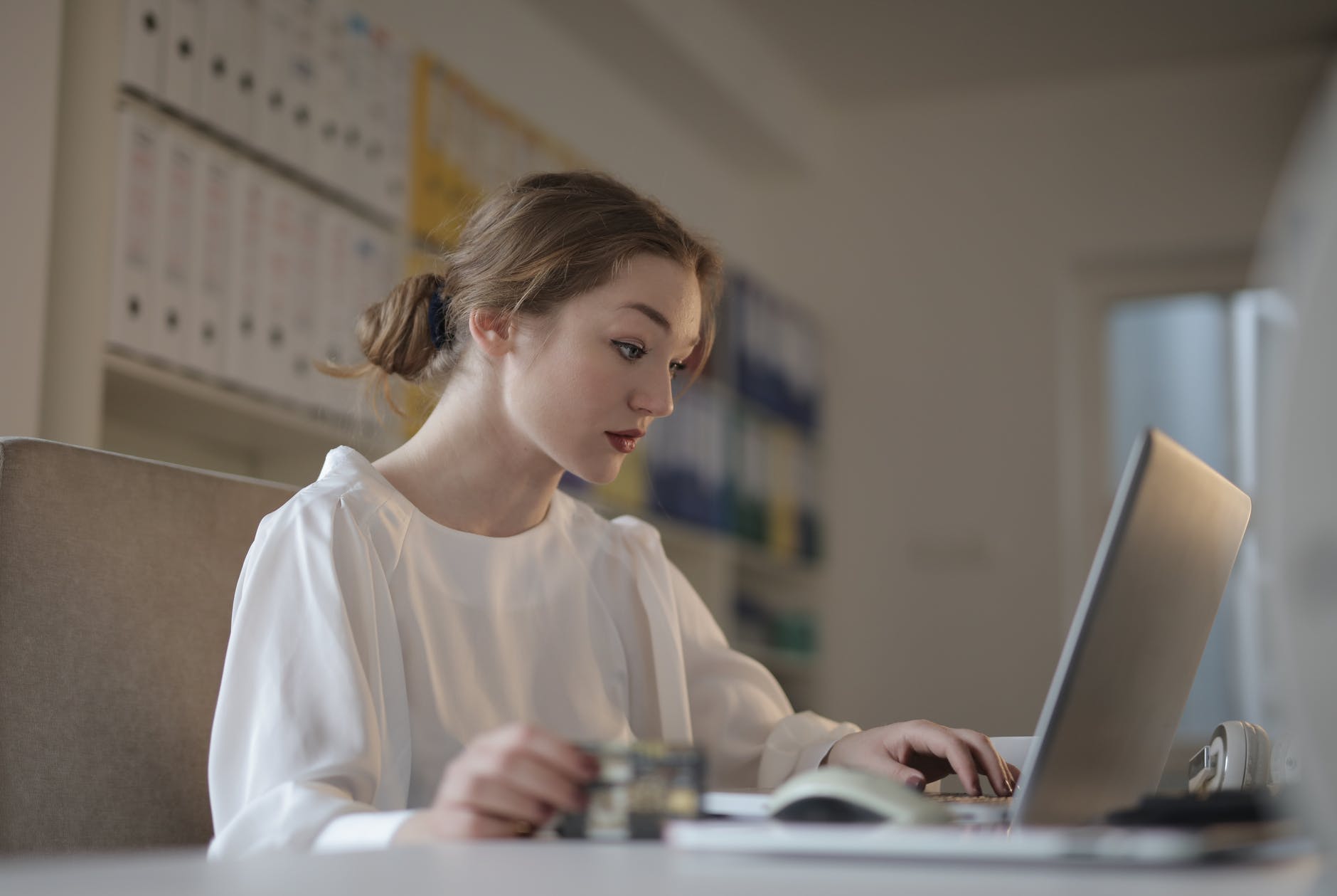 Woman with brown hair in bun and white blouse working on laptop.