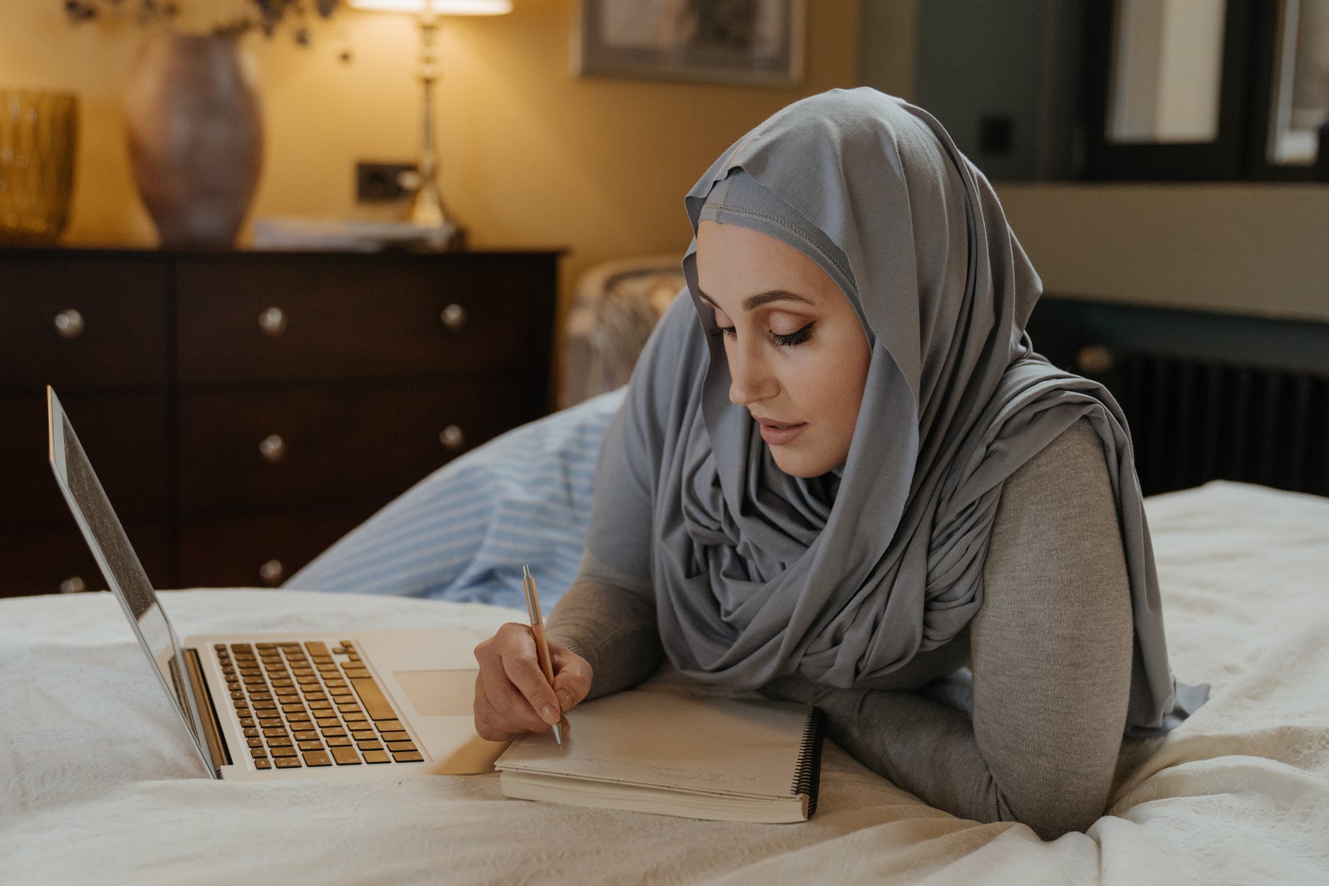 Woman with head dressing writing in notepad with open computer on bed.