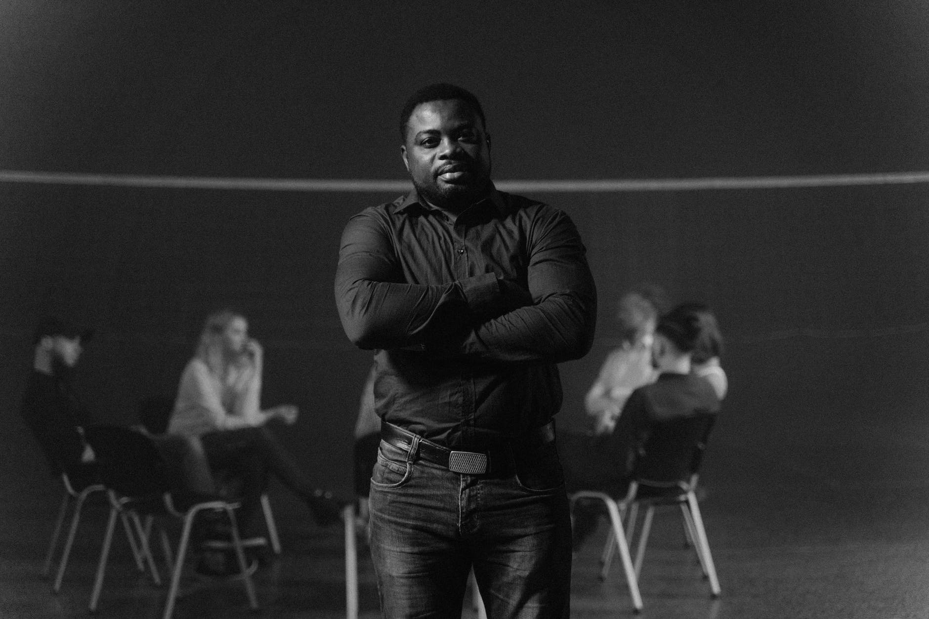 Black and white photograph. Black man with arms crossed and button up shirt. In the background, people in wheel chairs discuss.