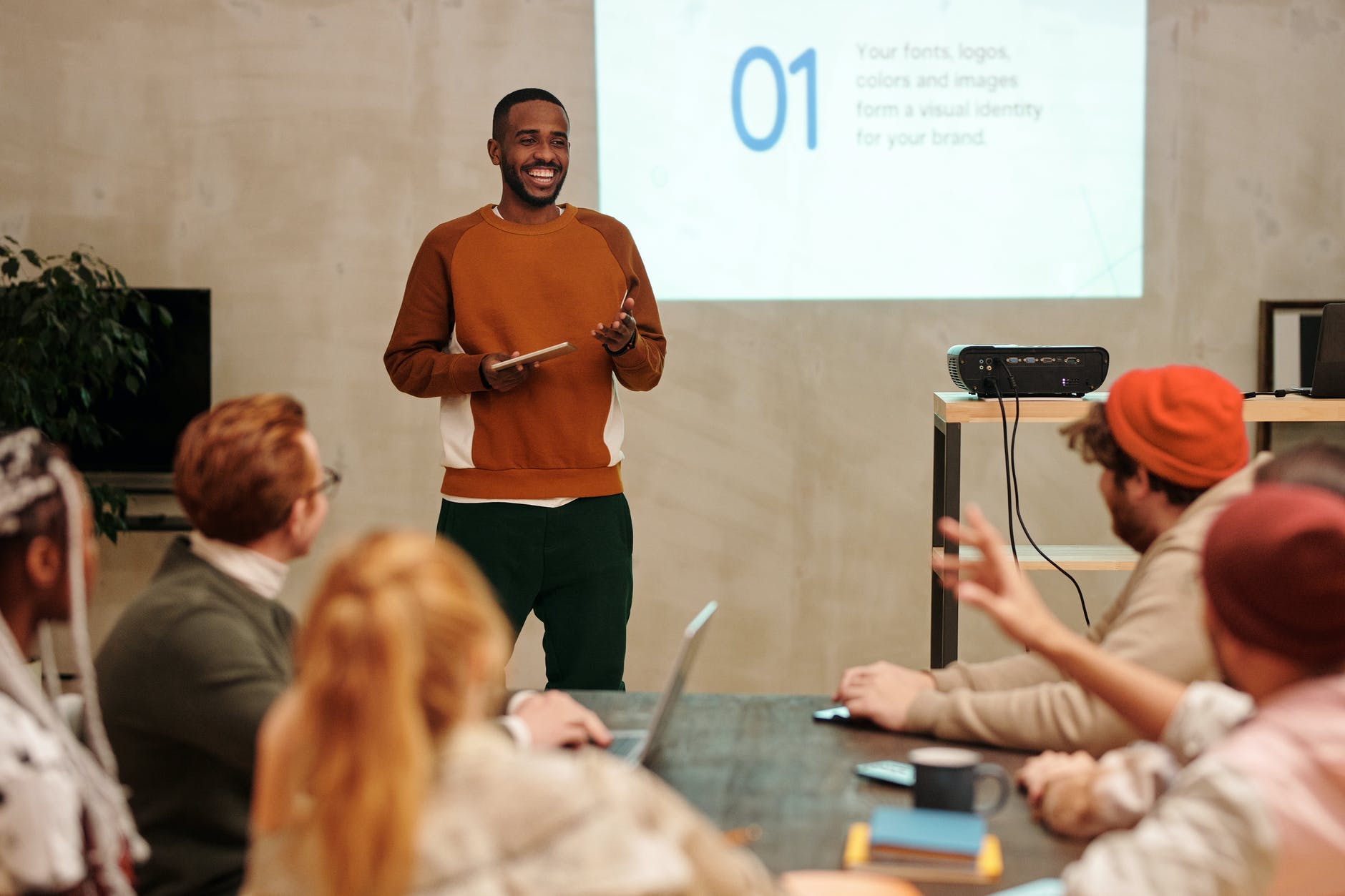 Black man laughing and presenting before four blurred out people seated around table.