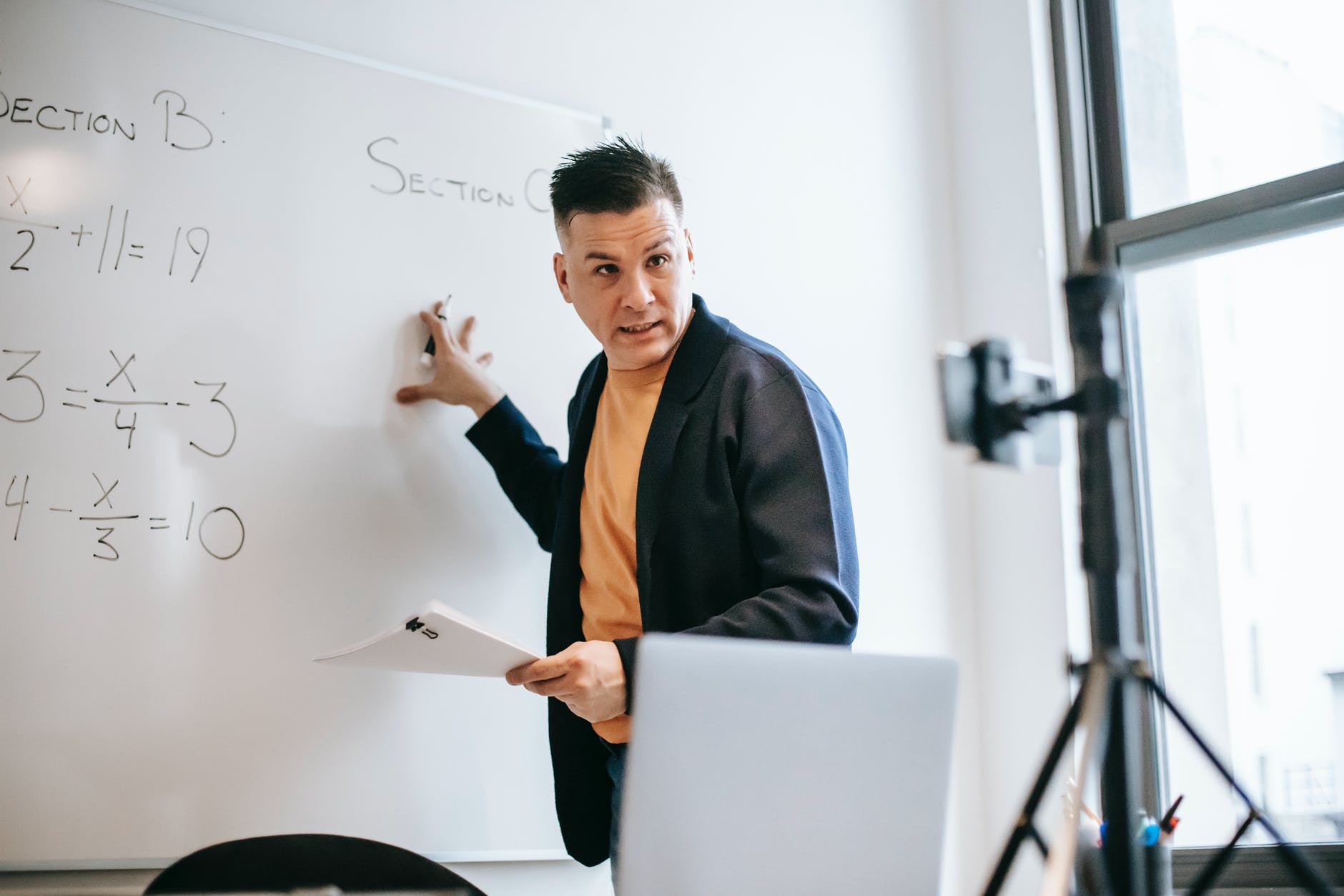 Male instructor working at whiteboard and holding paper with binder clip. A camera can be seen along with tripod.