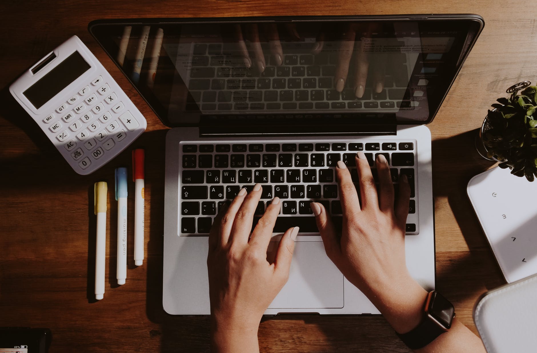 Laptop with hands on keyboard. Calculator and pens on desk.