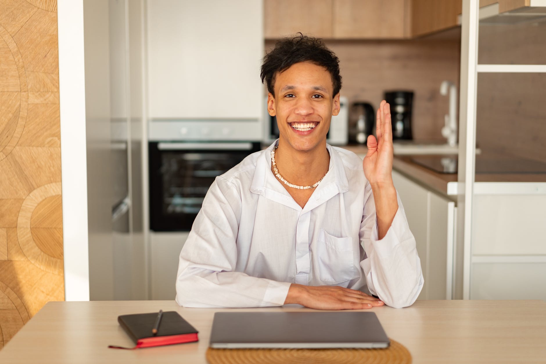 Person at table with one hand on table and the other elevated. The table has a laptop and black and red notebook. The person is smiling.