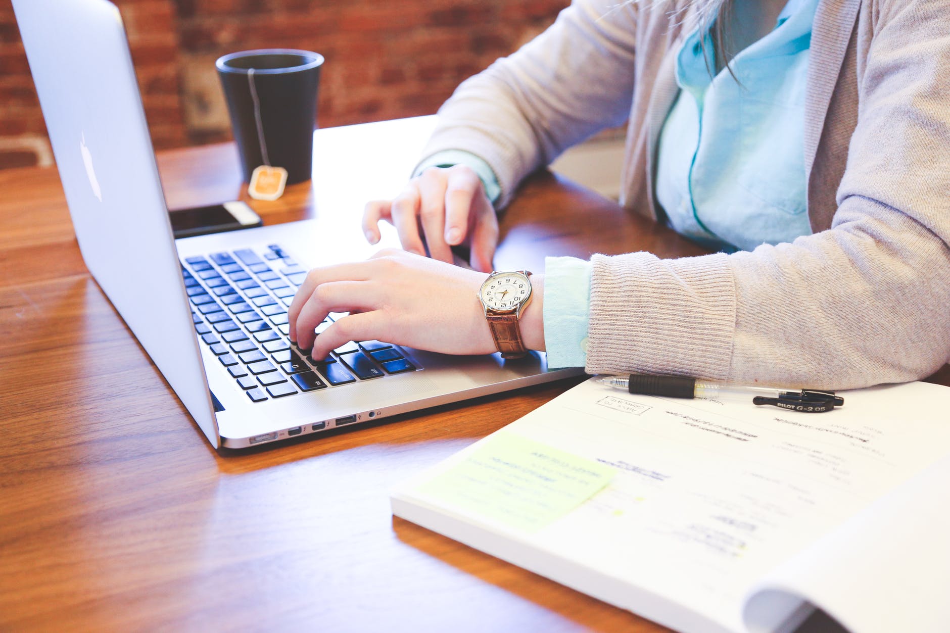 Two hands on a keyboard. A watch is visible on the left wrist. No head is visible. There is a tea cup on the right and a cell phone.