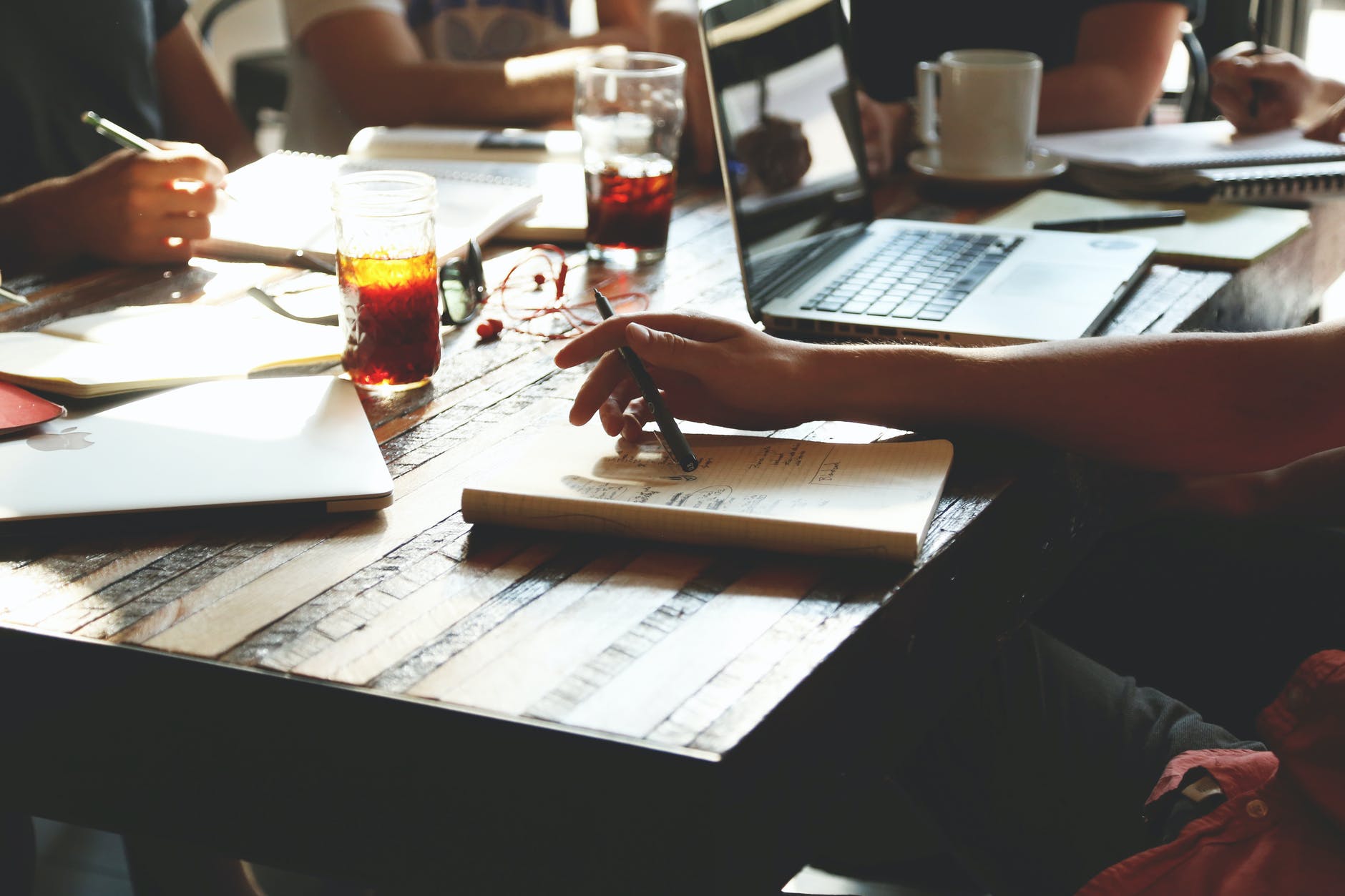 Table with laptop, notepads, and drinks. Arms of three different people are visible.