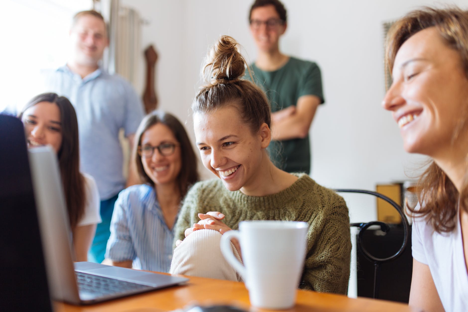 Four women seated smiling and looking at laptop that is open on the table and facing women. Two people are blurred in the background.
