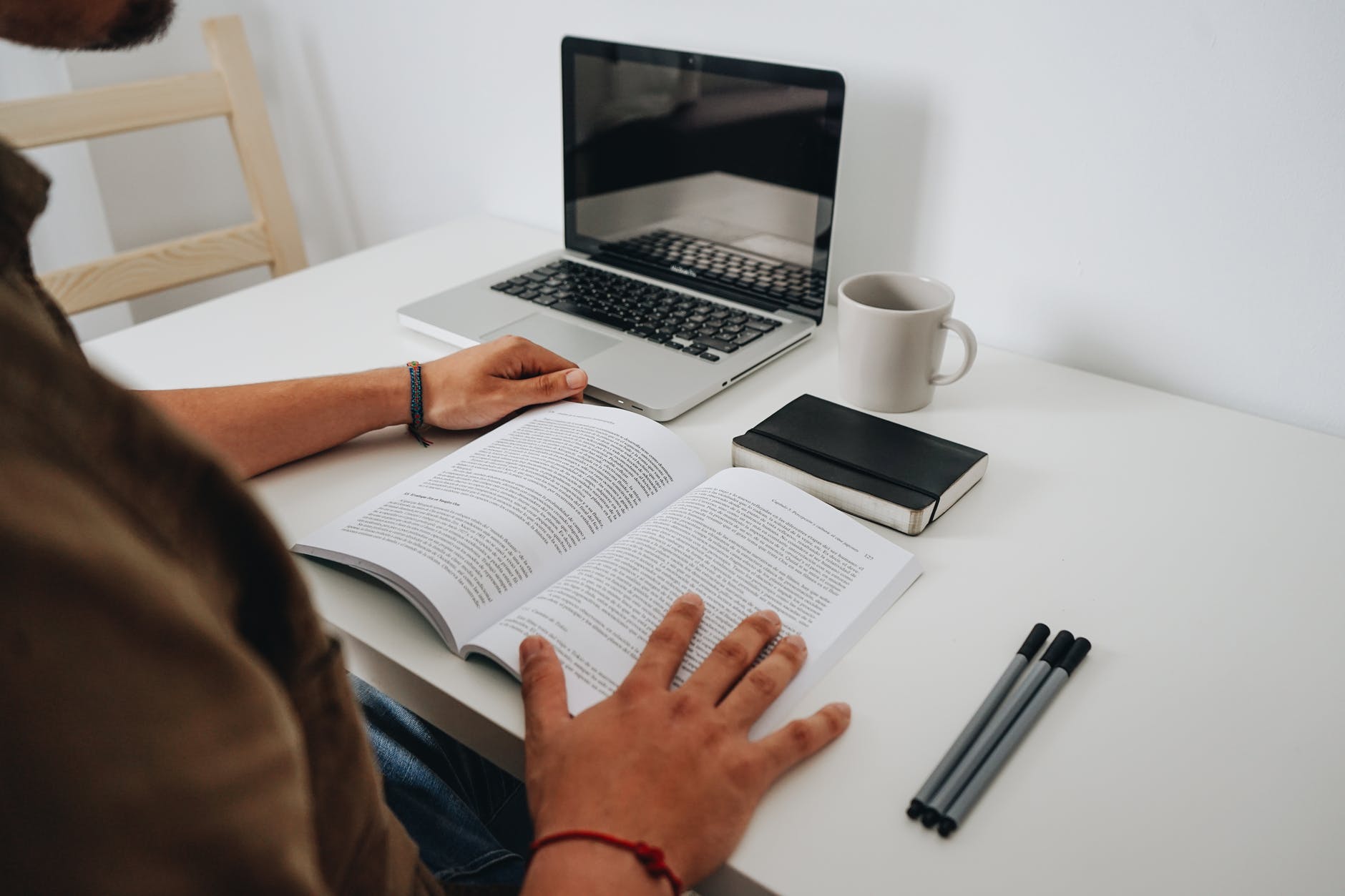 Desk with laptop, three pens, and open book. Two hands are holding book.