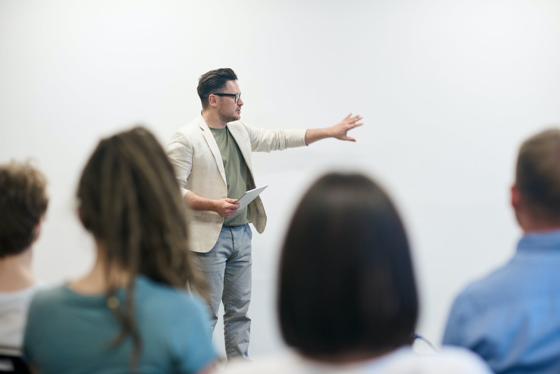 Male professor wearing white sports coat and jeans and holding notepad. Professor is wearing glasses and pointing at whiteboard. The backs of students are visible.