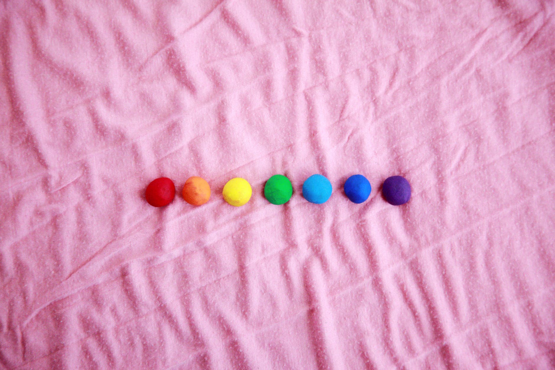 Pink blanket with balls of clay. From left to right: red, orange, yellow, green, light blue, dark blue, and purple. The balls are all lined up horizontally.
