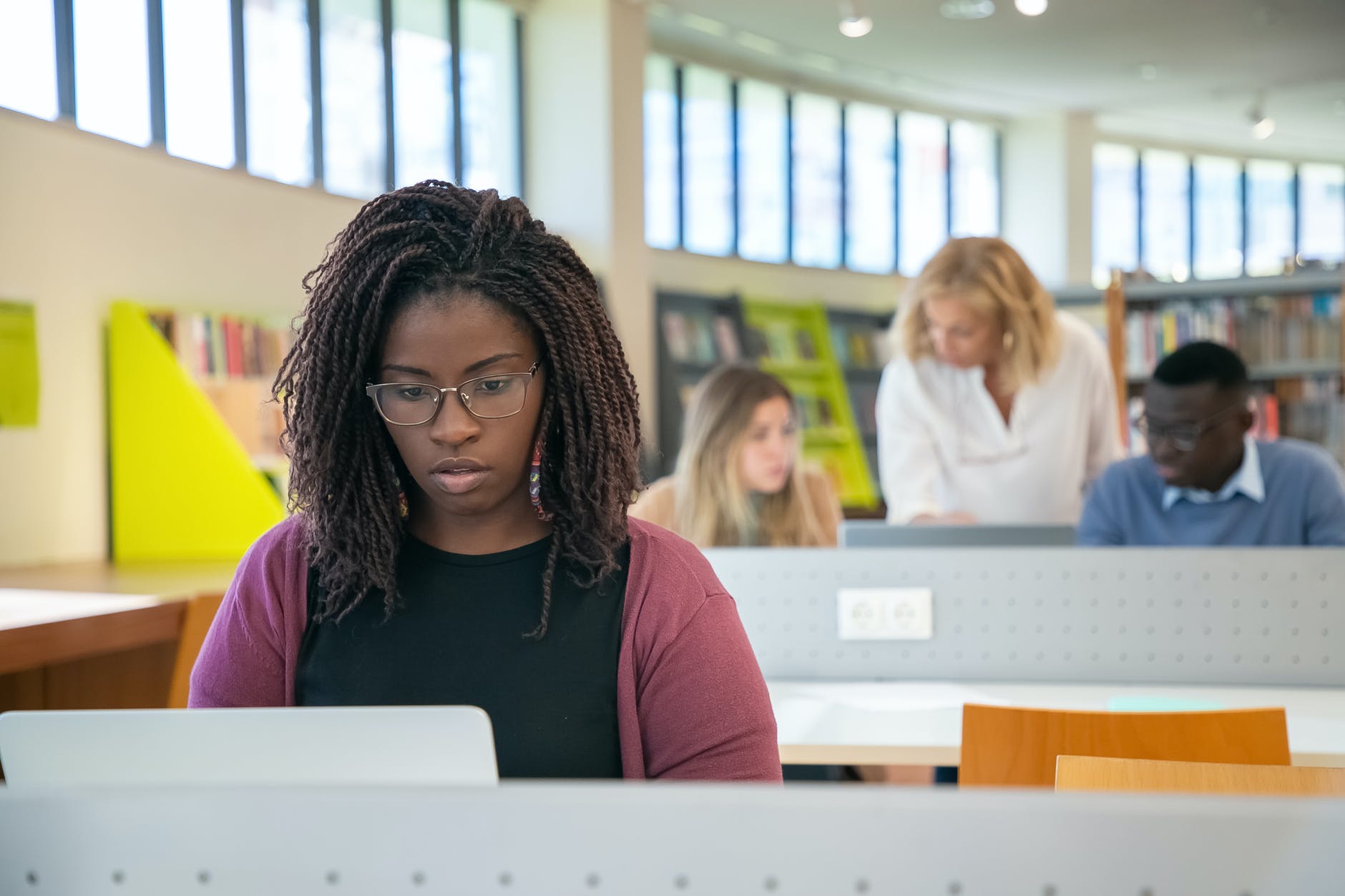 Black woman at keyboard of laptop in library. Books and desks are behind them. Faded: three people working at desk. 