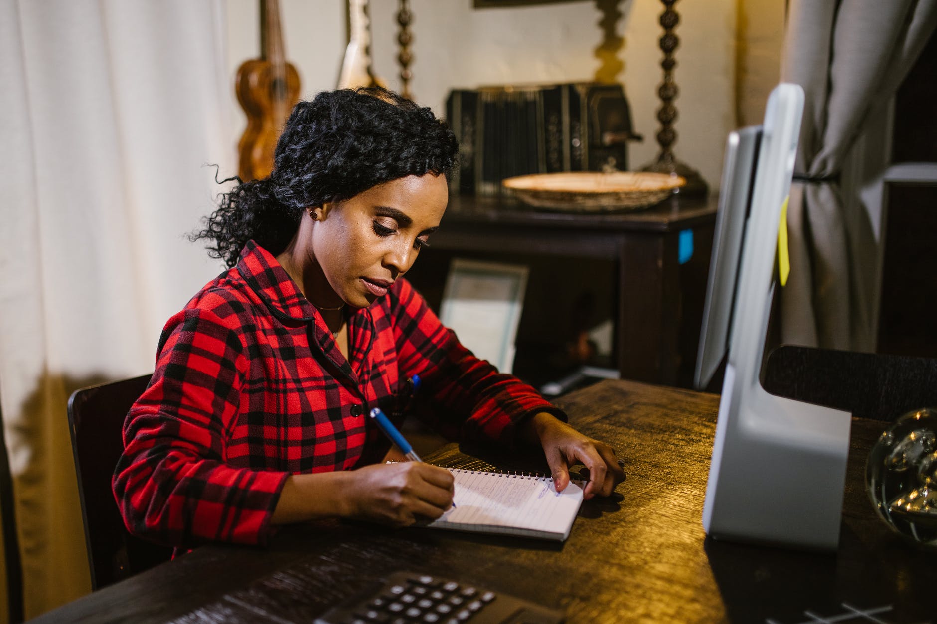 Black woman with curly hair writing in notebook in front of screen.