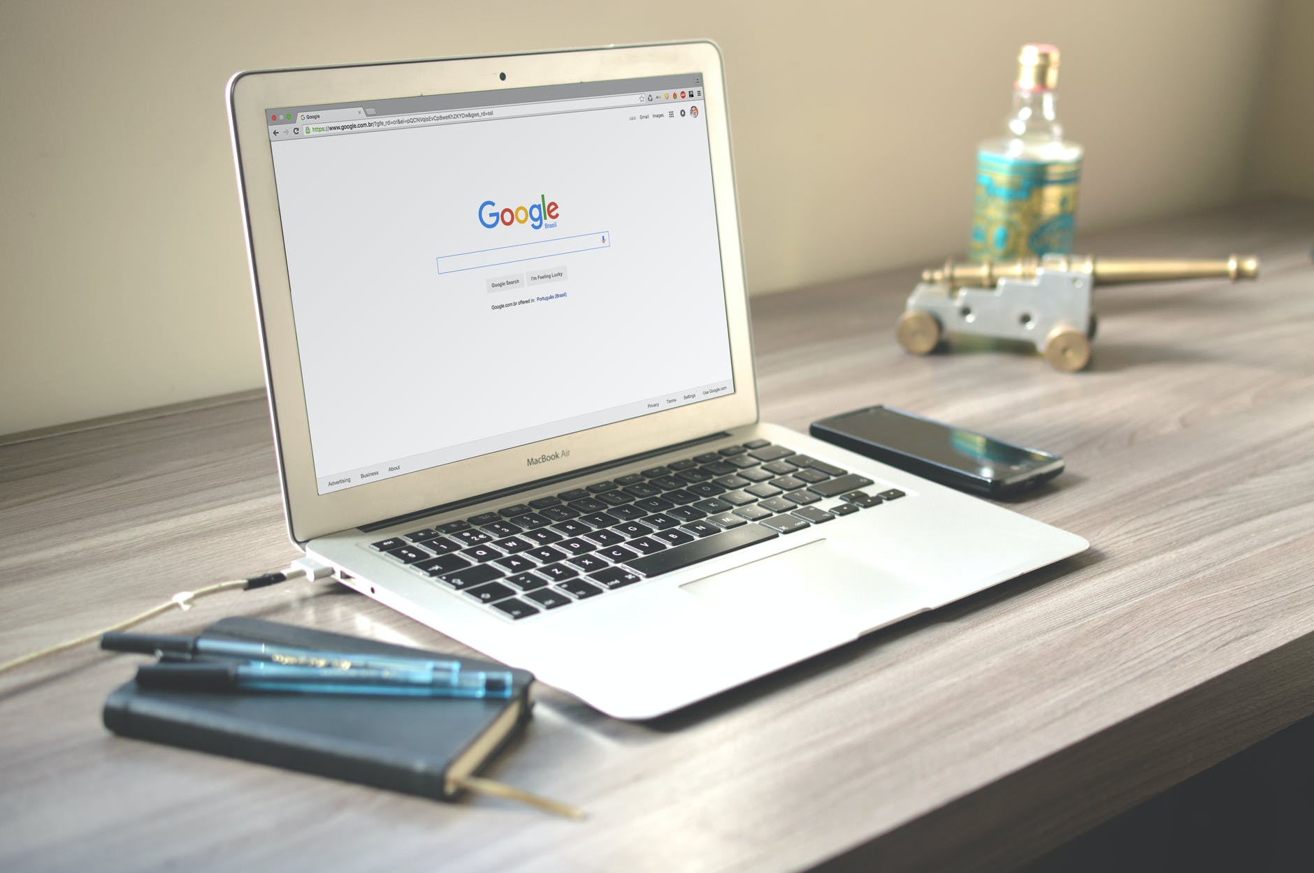 Laptop on desk with Google search homepage. The desk has a cell phone on the right of the laptop and a notebook and two pens on the left.