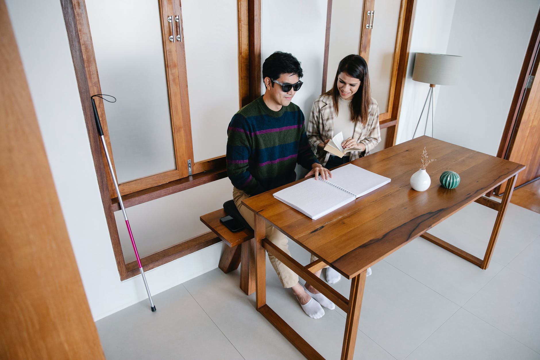 Man wearing dark glasses seated at table next to cane and woman helping read book.