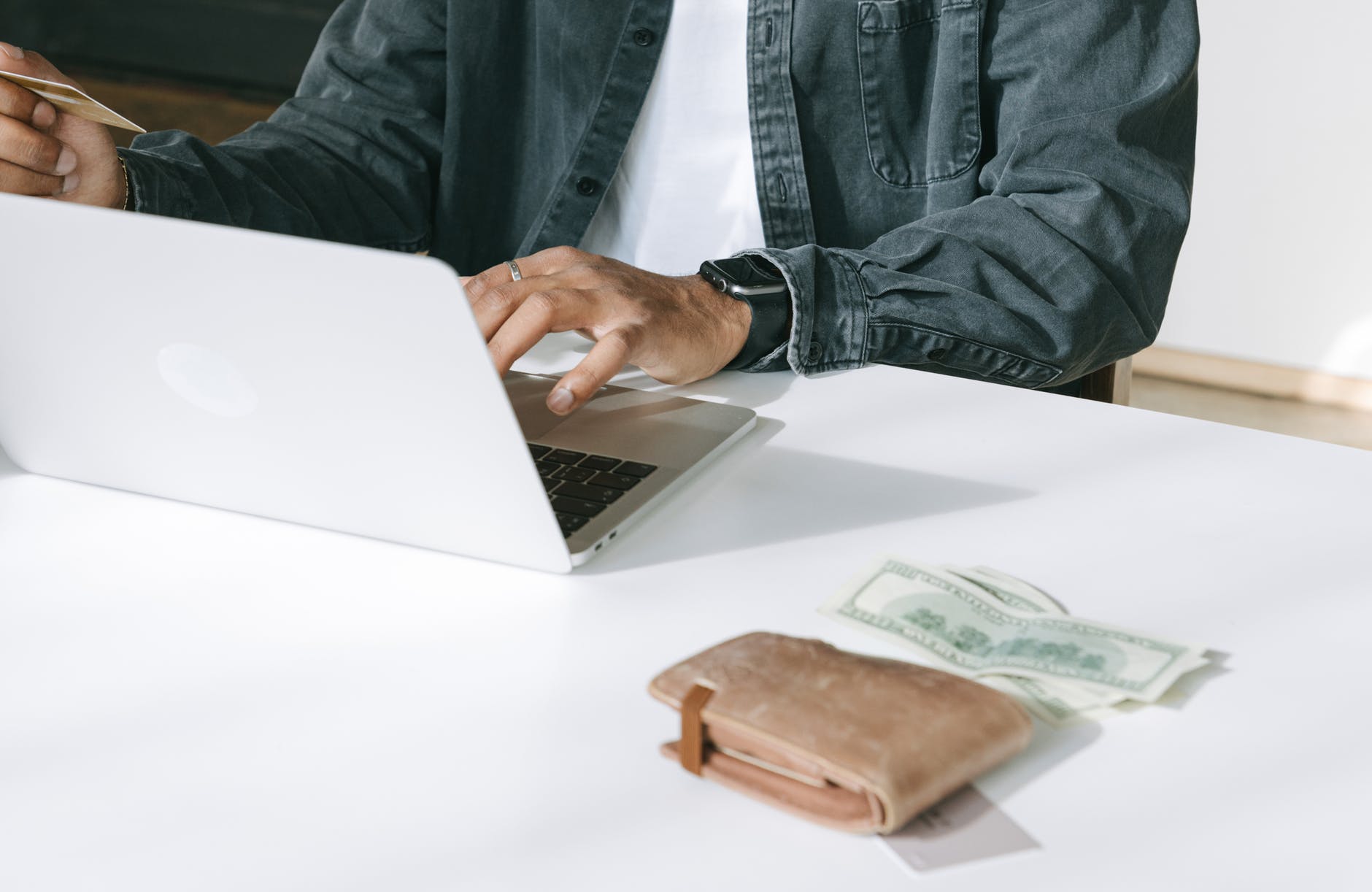 White table with laptop and hand typing. There is a wallet and money on the table.