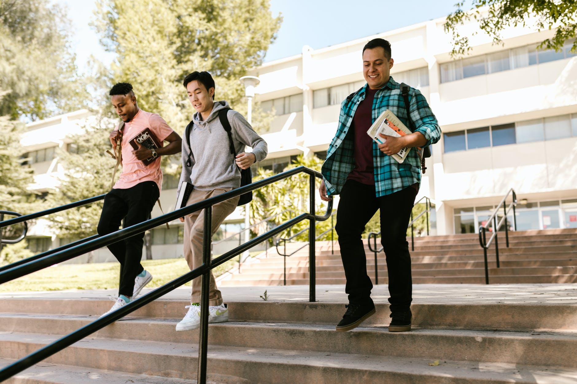 Left to right: black man holding book, asian man holding book and wearing hoodie and man holding notepads all walking downstairs.