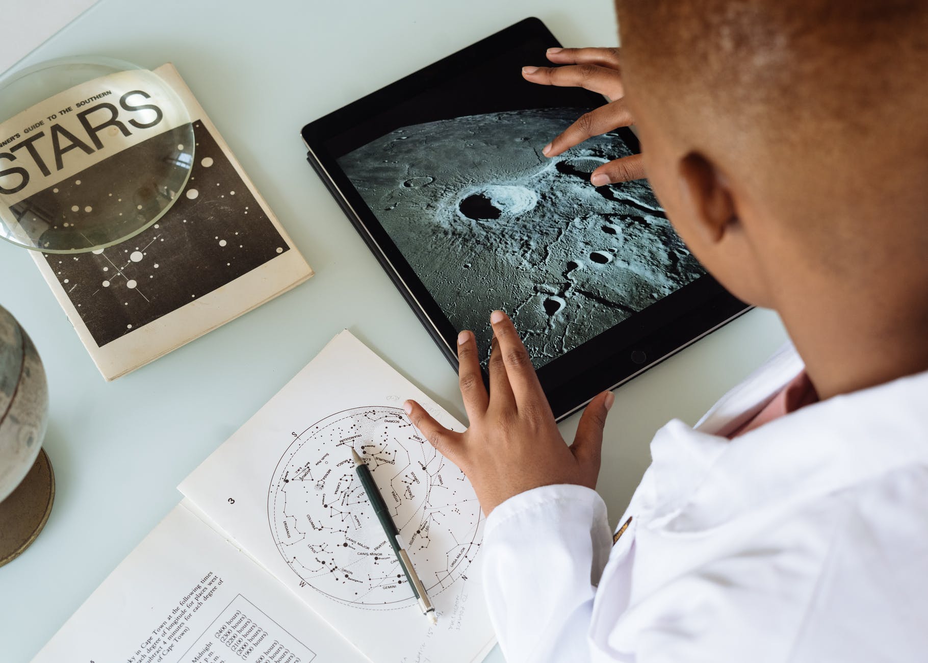 iPad with moon crater on screen. Black hands and back of head of person using the iPad. A book on starts is on the table.