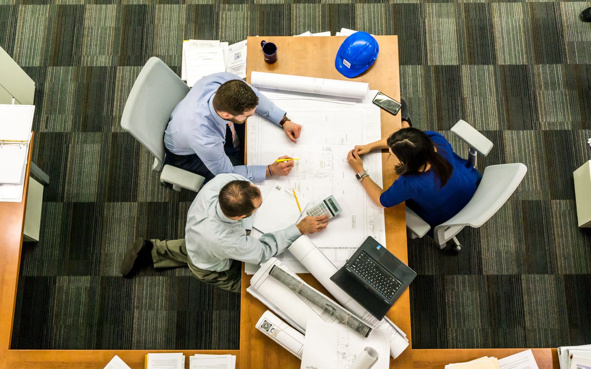 Top down view of group of three people working on plans with calculator, highlighter, laptop, and cell phone.