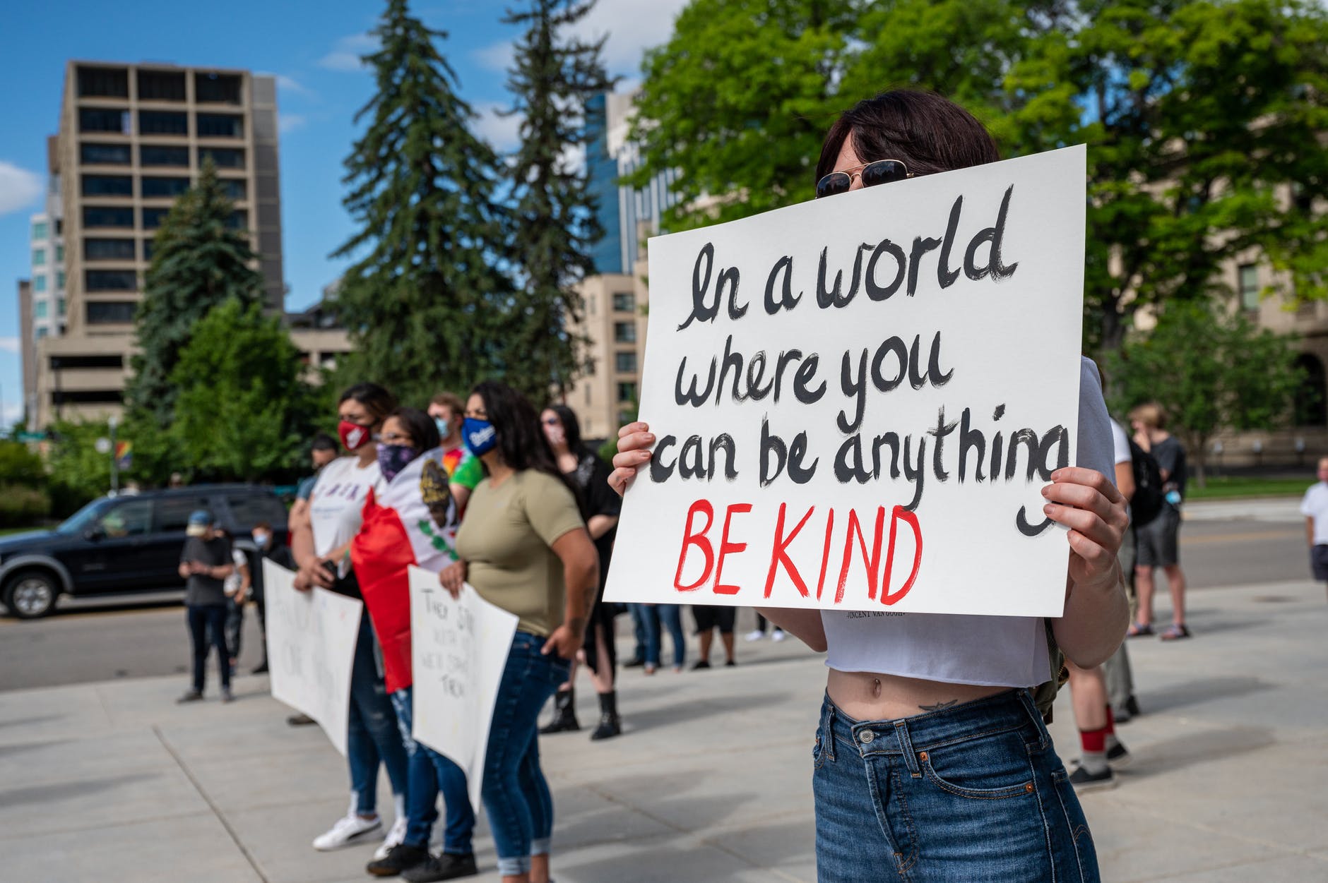 Protesters wearing masks and holding a sign "In a world where you can be anything, be kind."