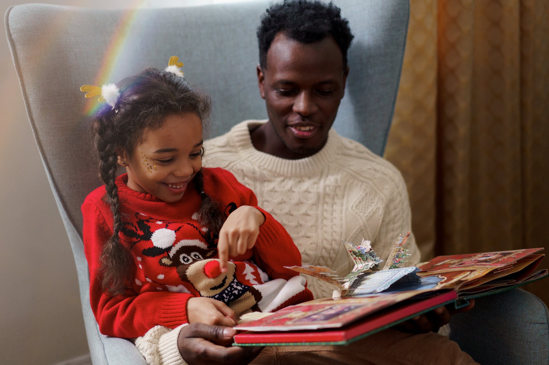 Black man and girl reading storybook. Both are wearing winter holiday sweaters.