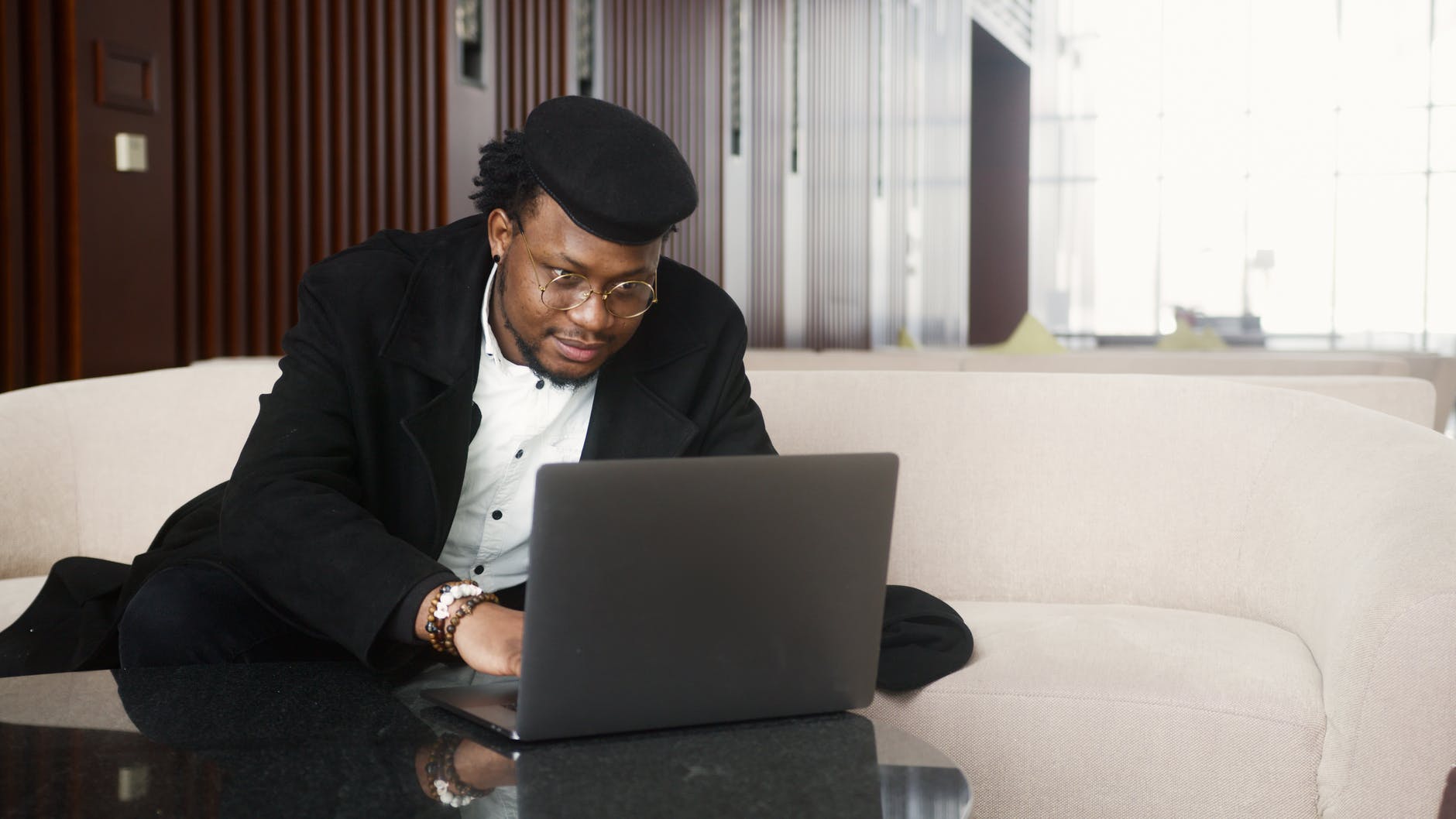 Black man on couch typing on laptop on table. He is wearing black coat.
