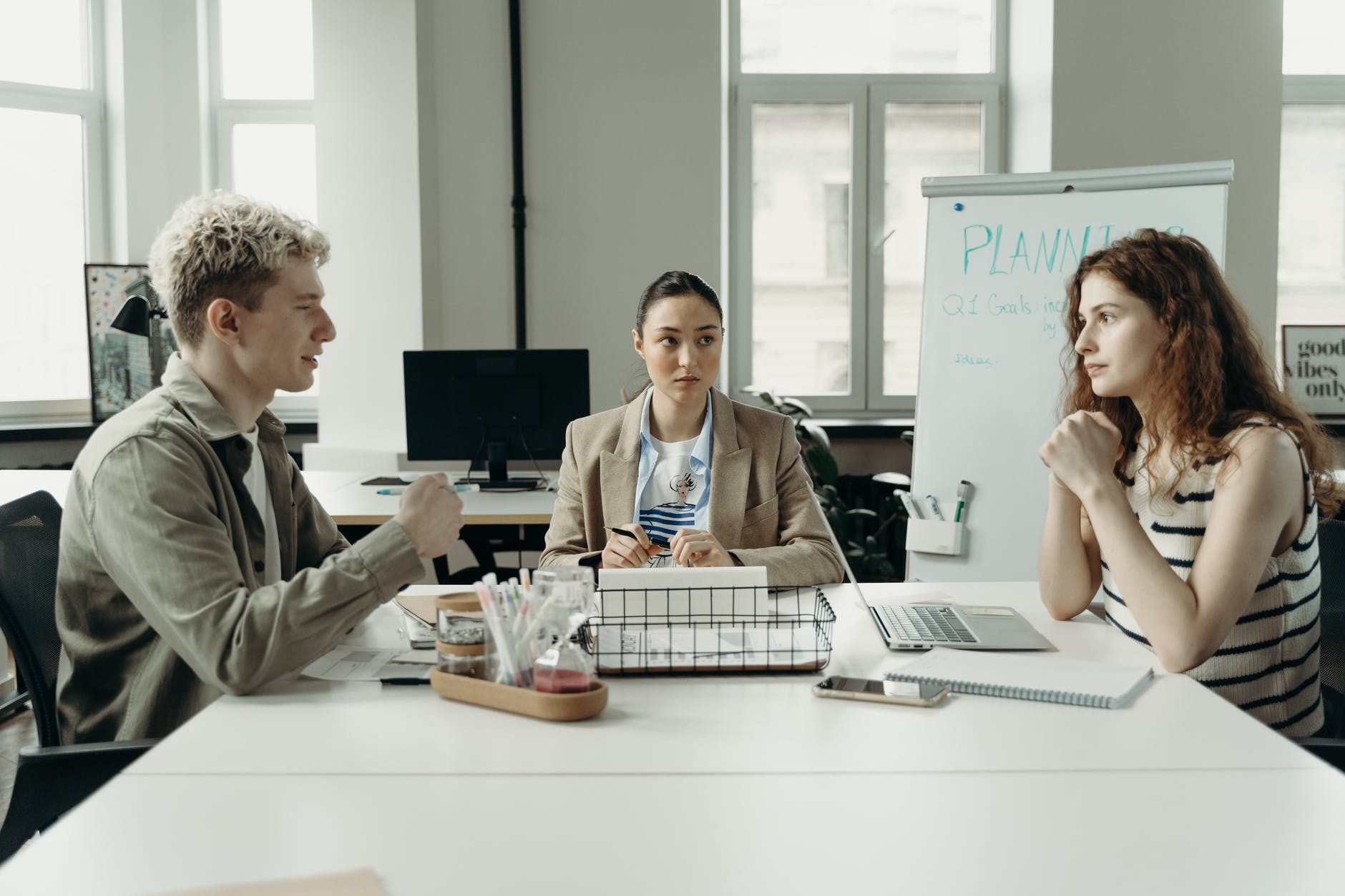 Three people seated around rectangular table. Left man with blond hair; center woman with pen; right woman with laptop.