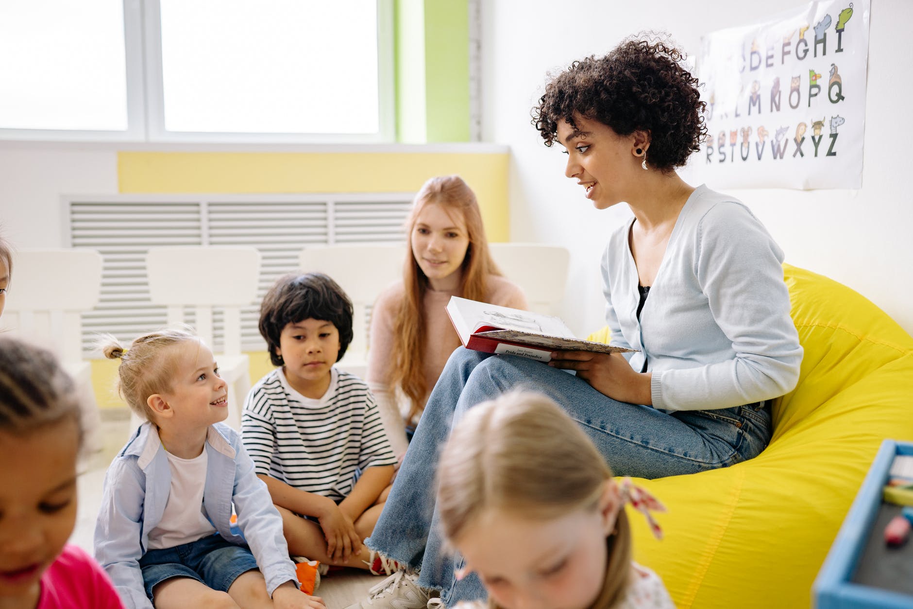 Woman with curly hair reading book to group of children that surround them.