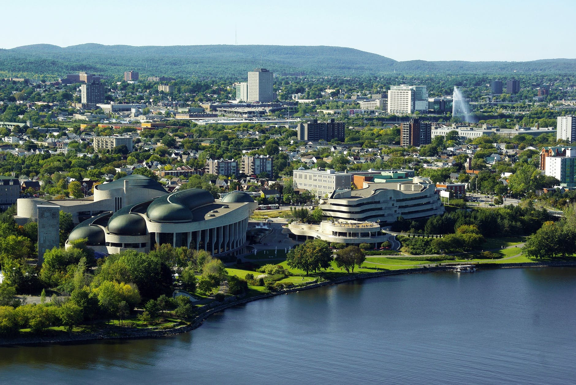 Aerial view of city with body of water and green lawns. Several modern buildings near the waterfront can be seen.