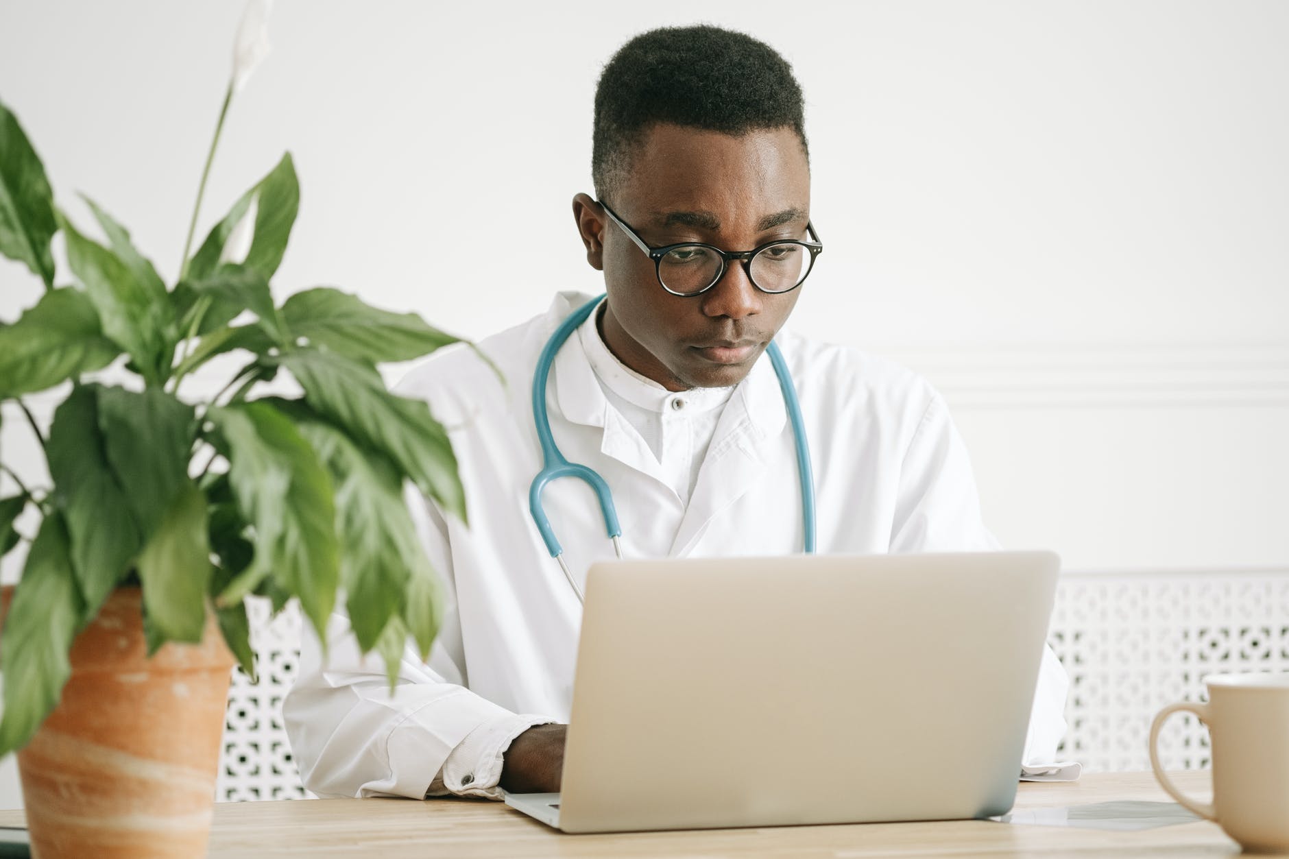 Black medical student with white lab coat and stethoscope working on laptop.