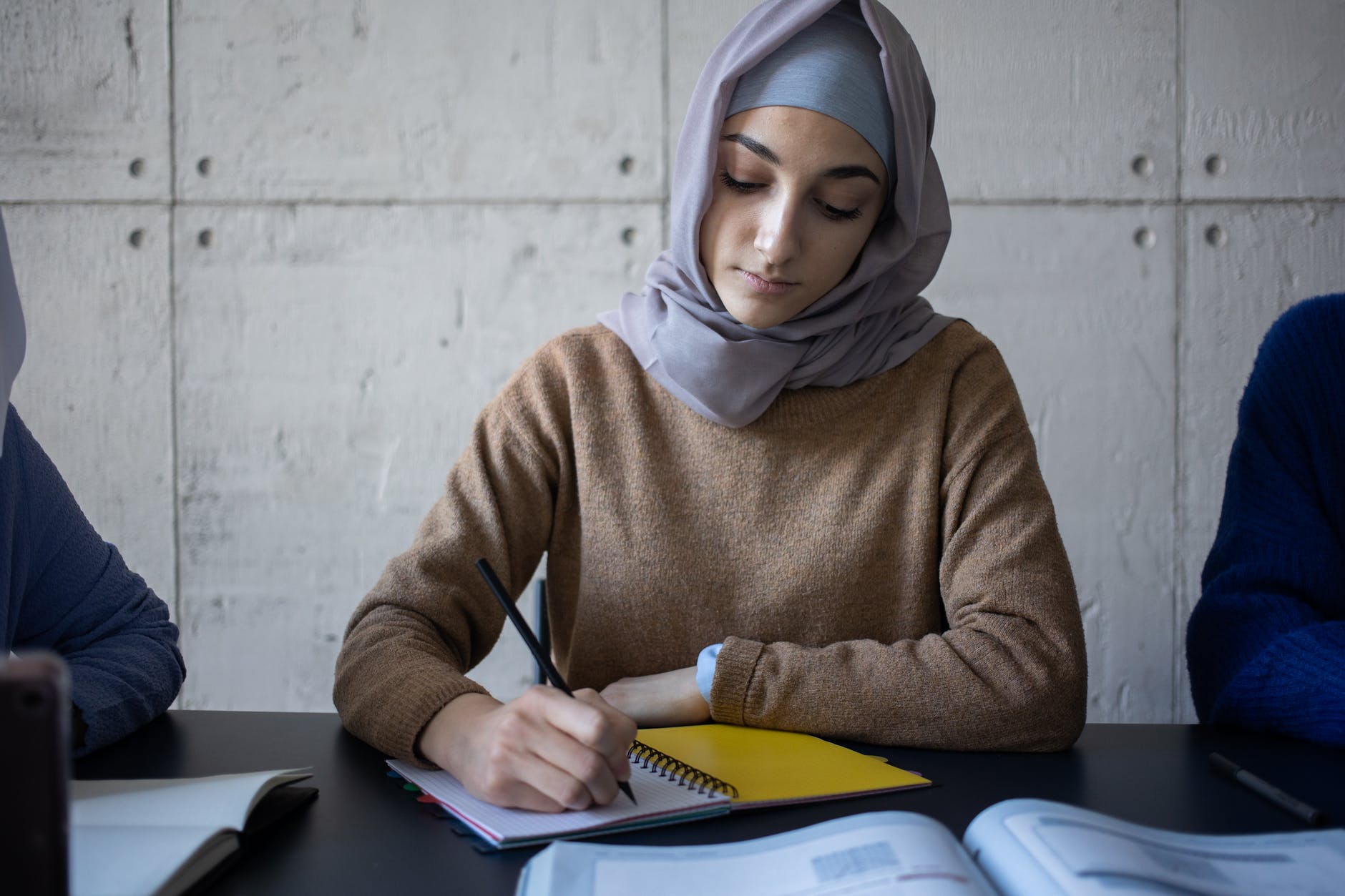 Woman taking notes. Wearing head scarf and brown sweater.