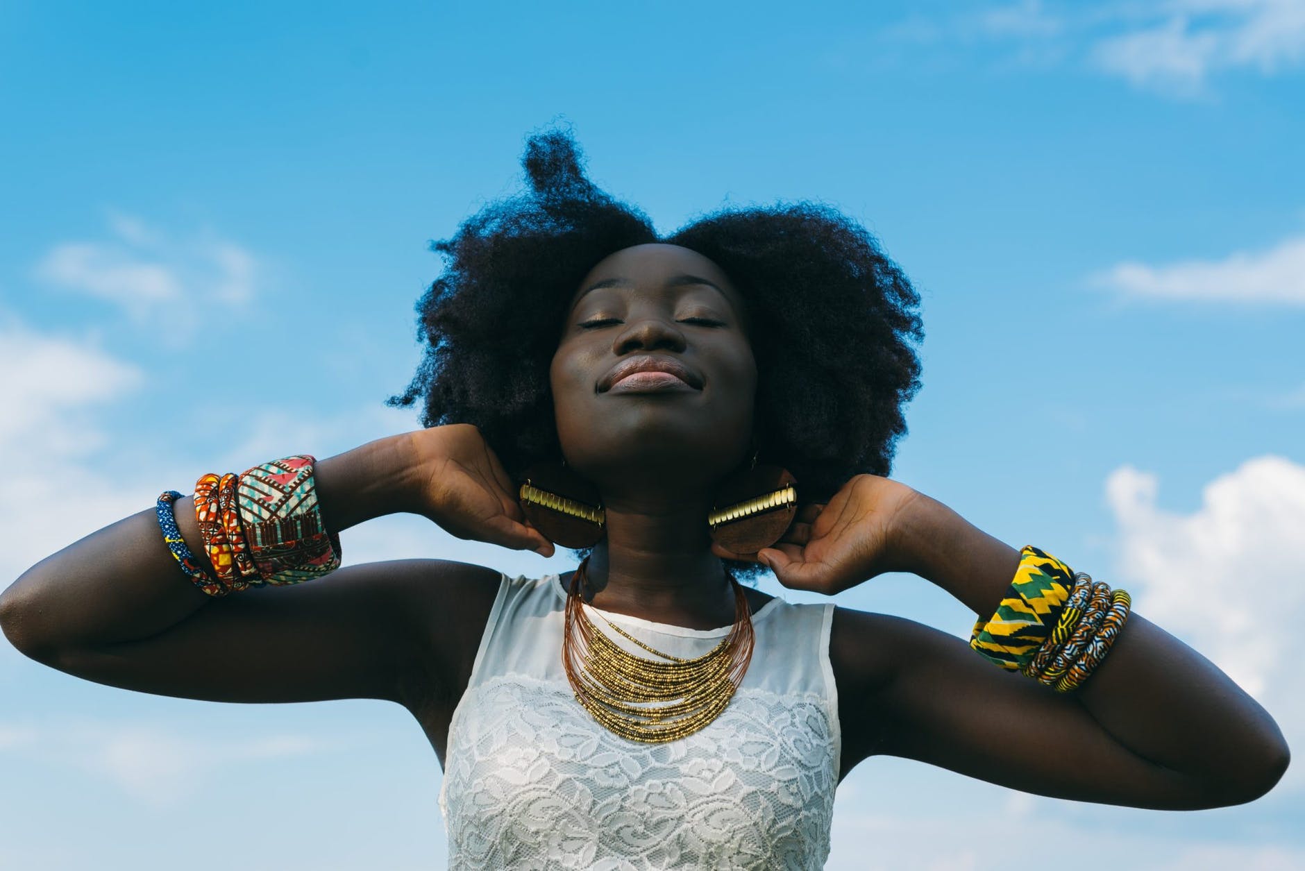 Black woman with colorful jewelry and eyes closed, hands behind neck, smiling.