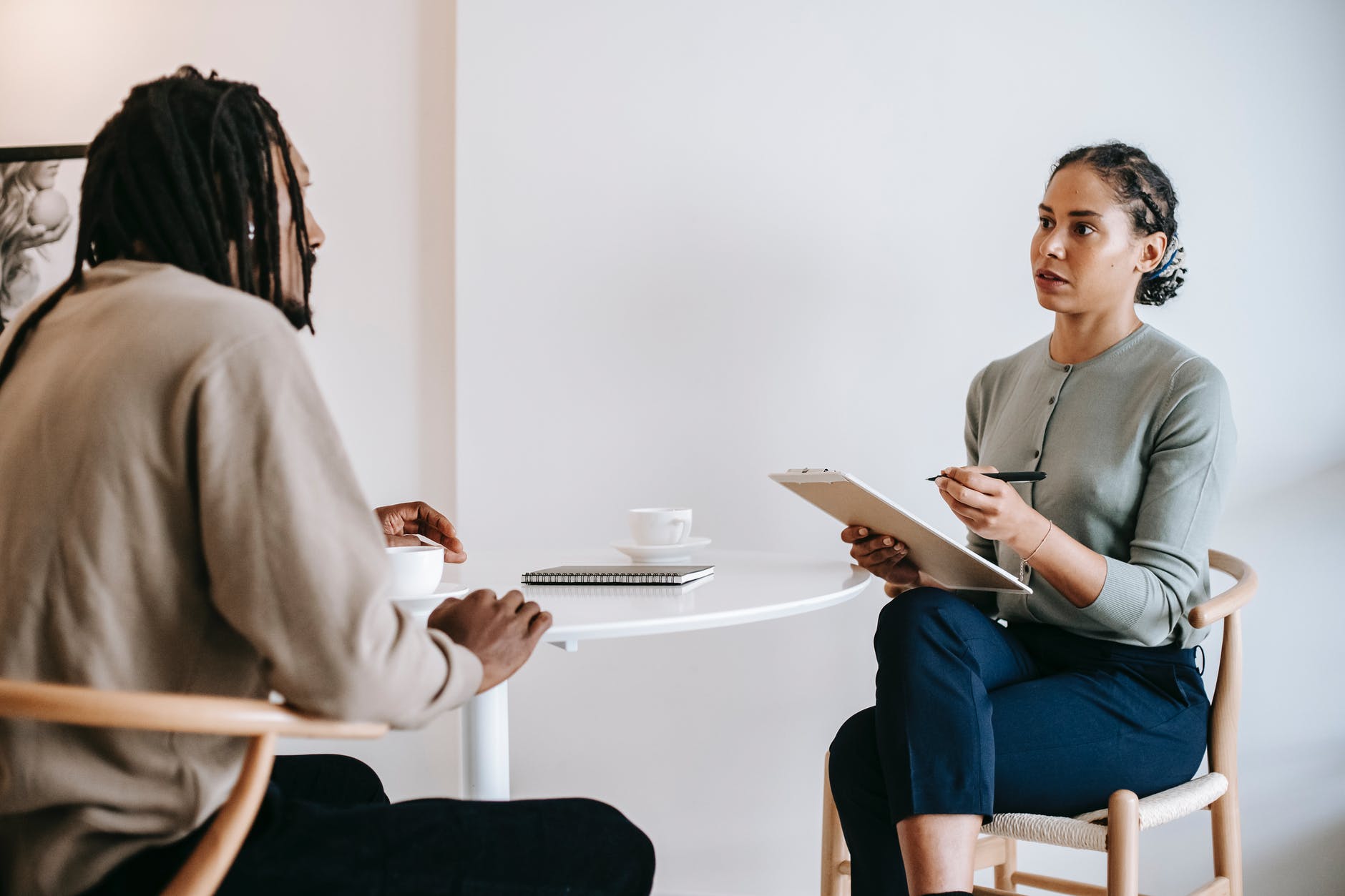 A black man being interviewed by a black woman with short ponytail and clipboard. There is a white round table with two white tea cups.