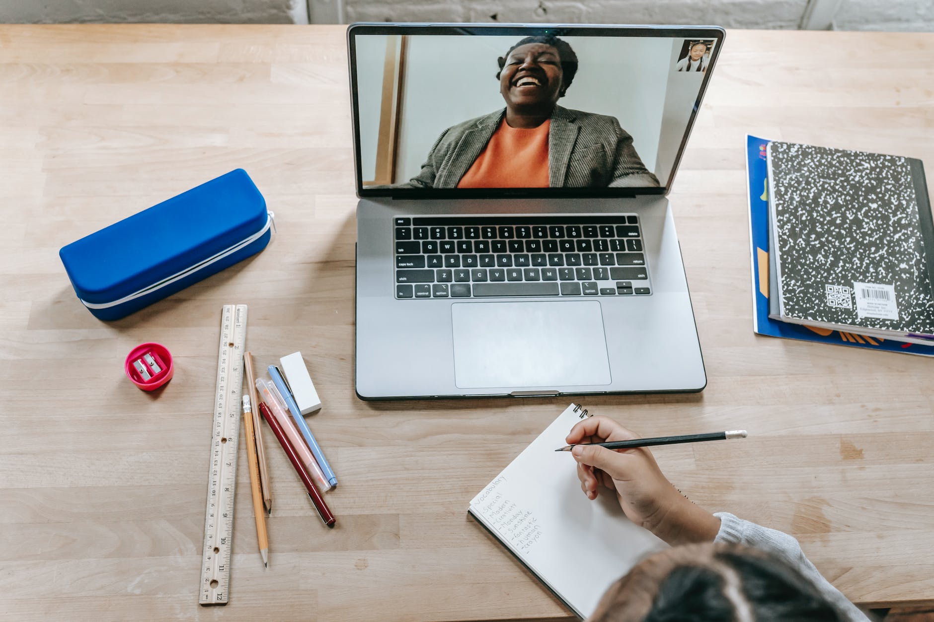 Top down view of laptop with laughing Black woman on screen. Hand near laptop is taking notes.