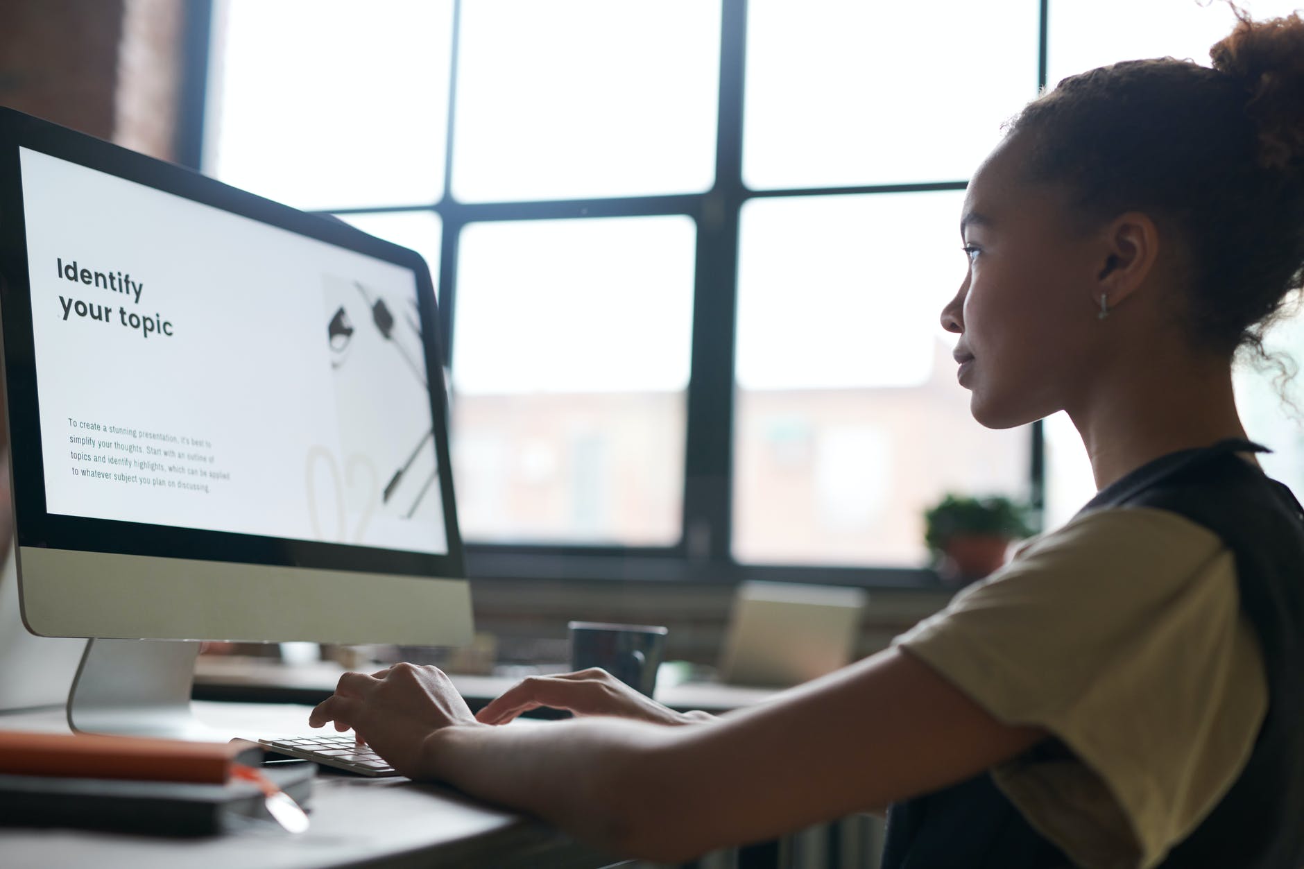 Woman working on desktop Mac computer with "Identify your topic" on screen.