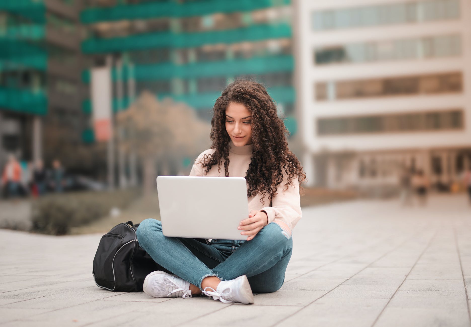 woman with curly brown hair sitting down with legs crossed and working on white laptop