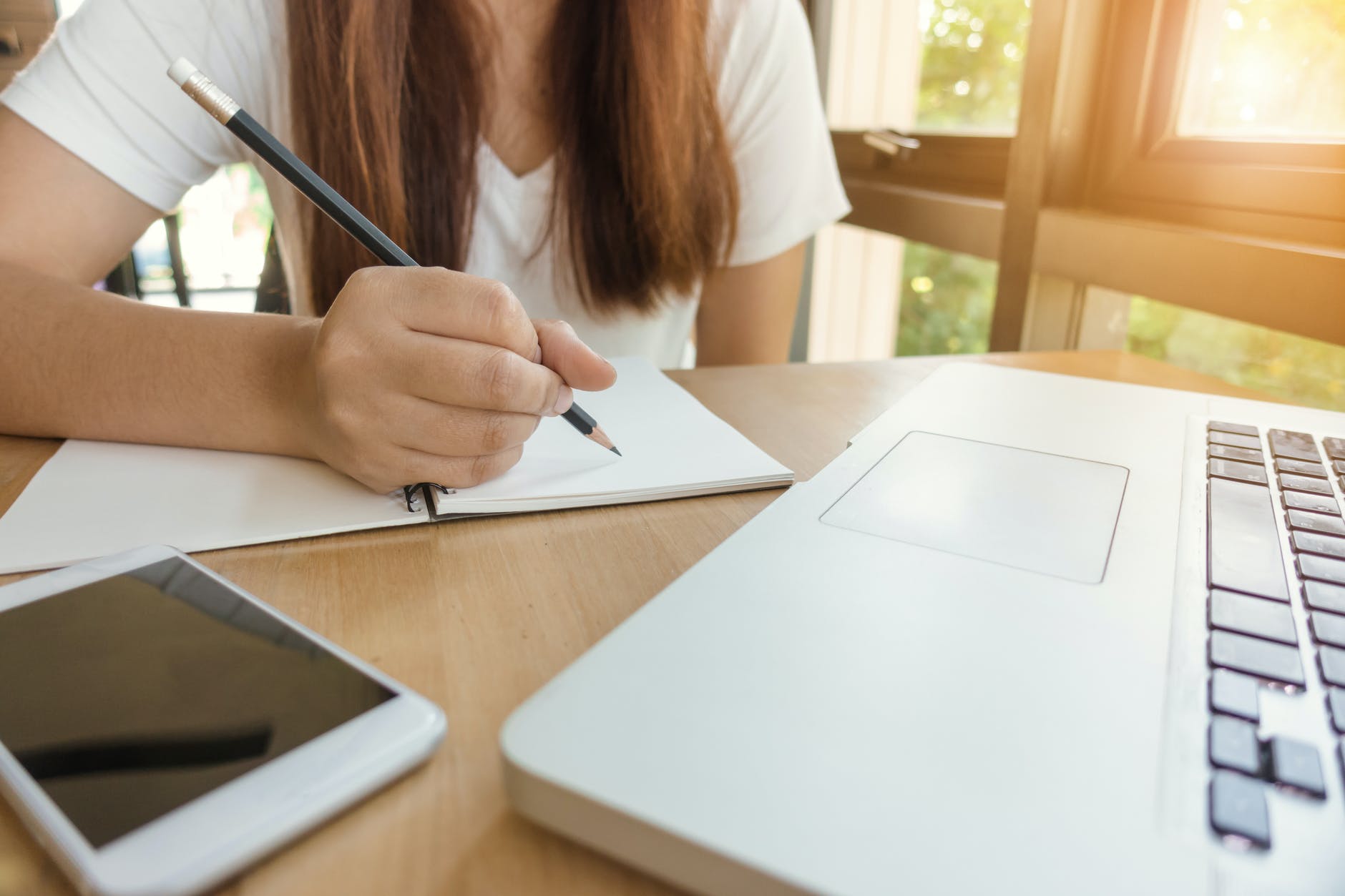 Person with long hair holding pencil and writing in notepad next to laptop