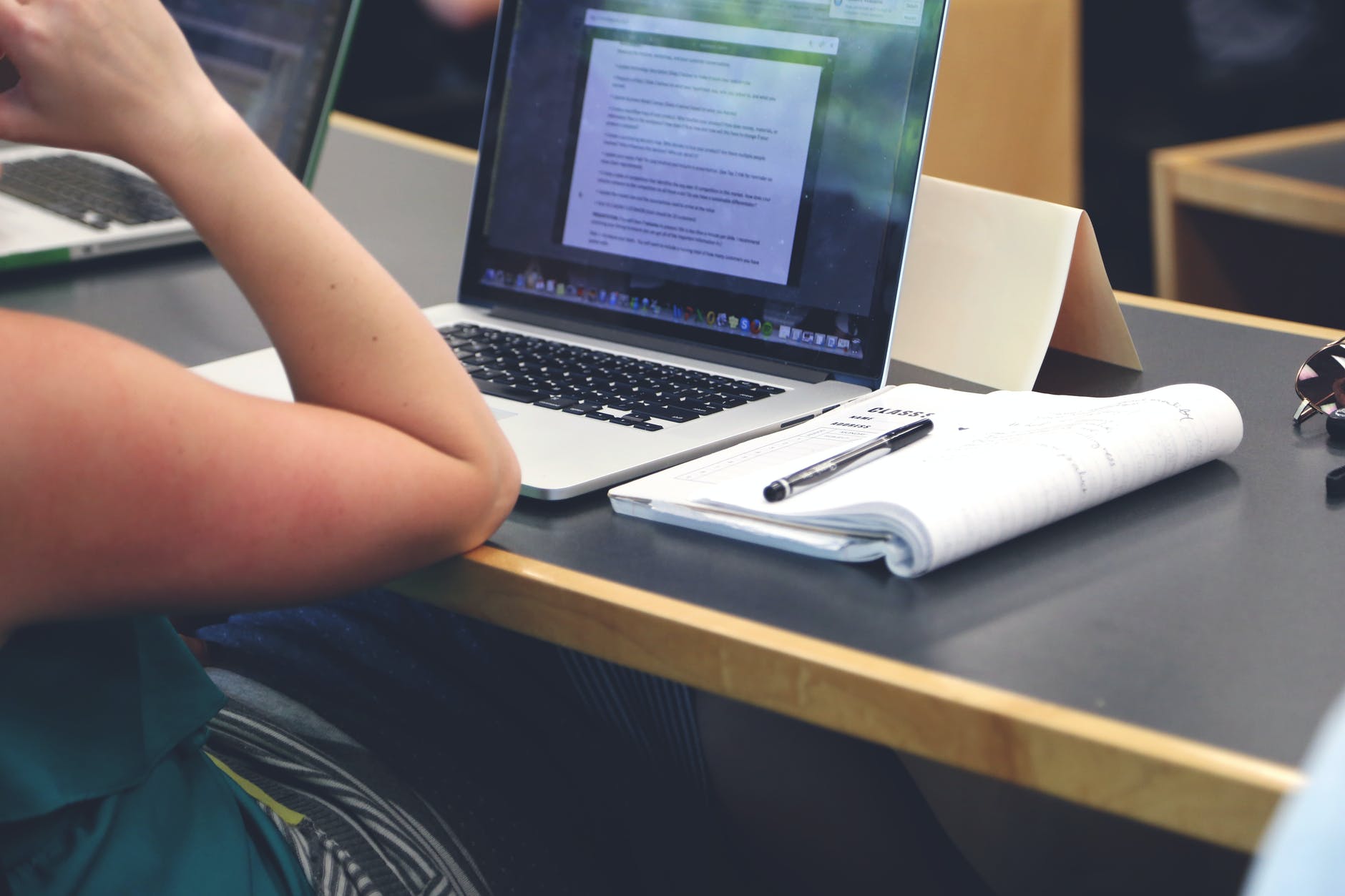 Desk with notebook and open laptop. Elbow of person sitting in front of laptop.