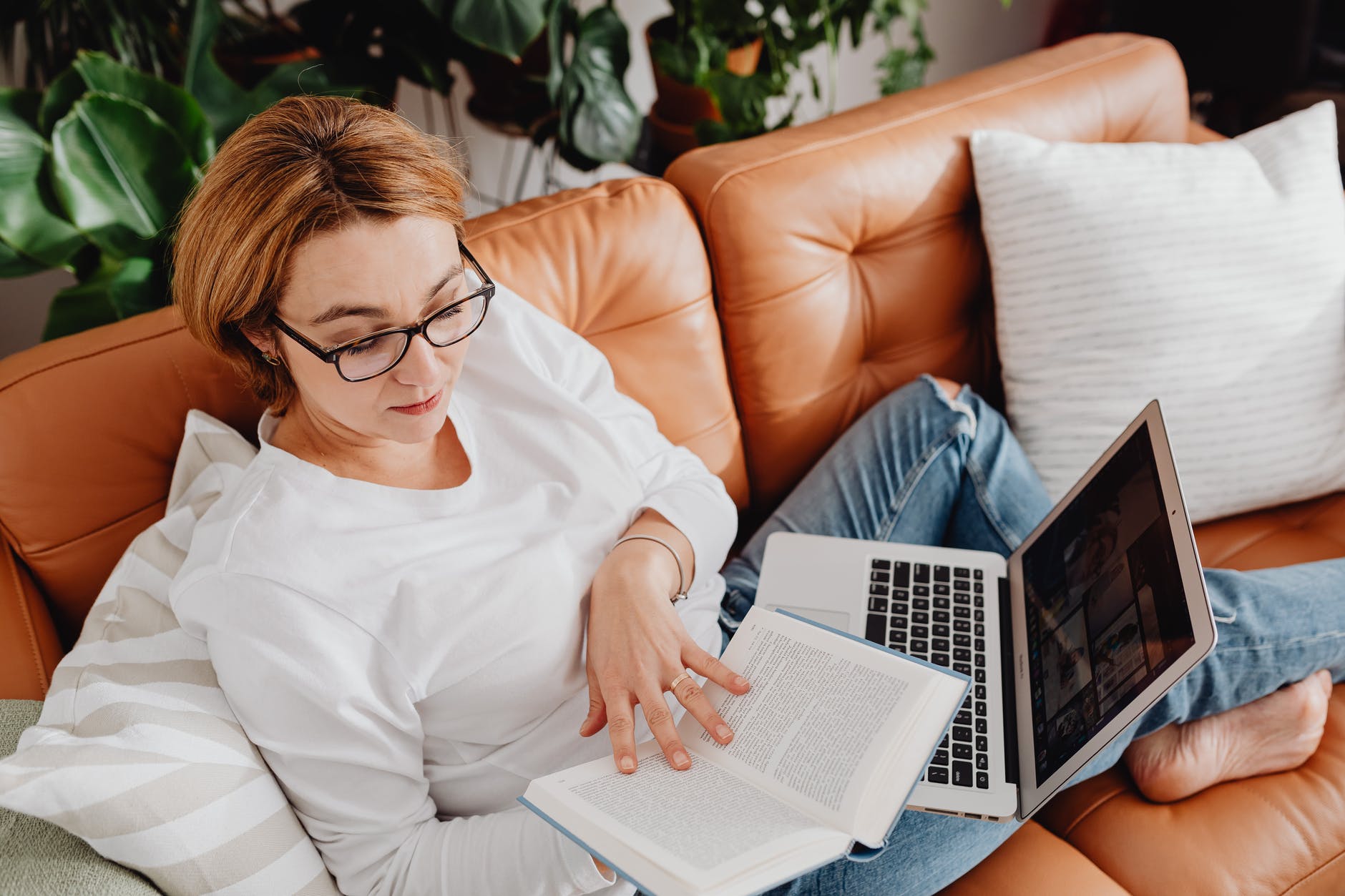 woman reading a book and using laptop on orange couch